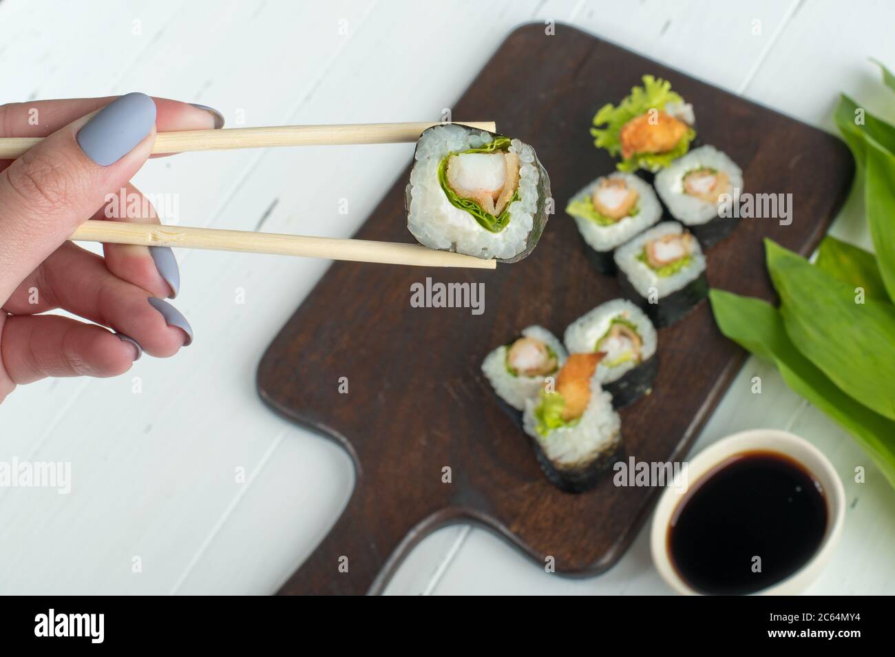 Ensemble de sushis avec crevettes frites et salade servi sur un plateau en bois sombre. Femme tenant main un rouleau de sushi frais avec des baguettes en bois. Fond gris Banque D'Images