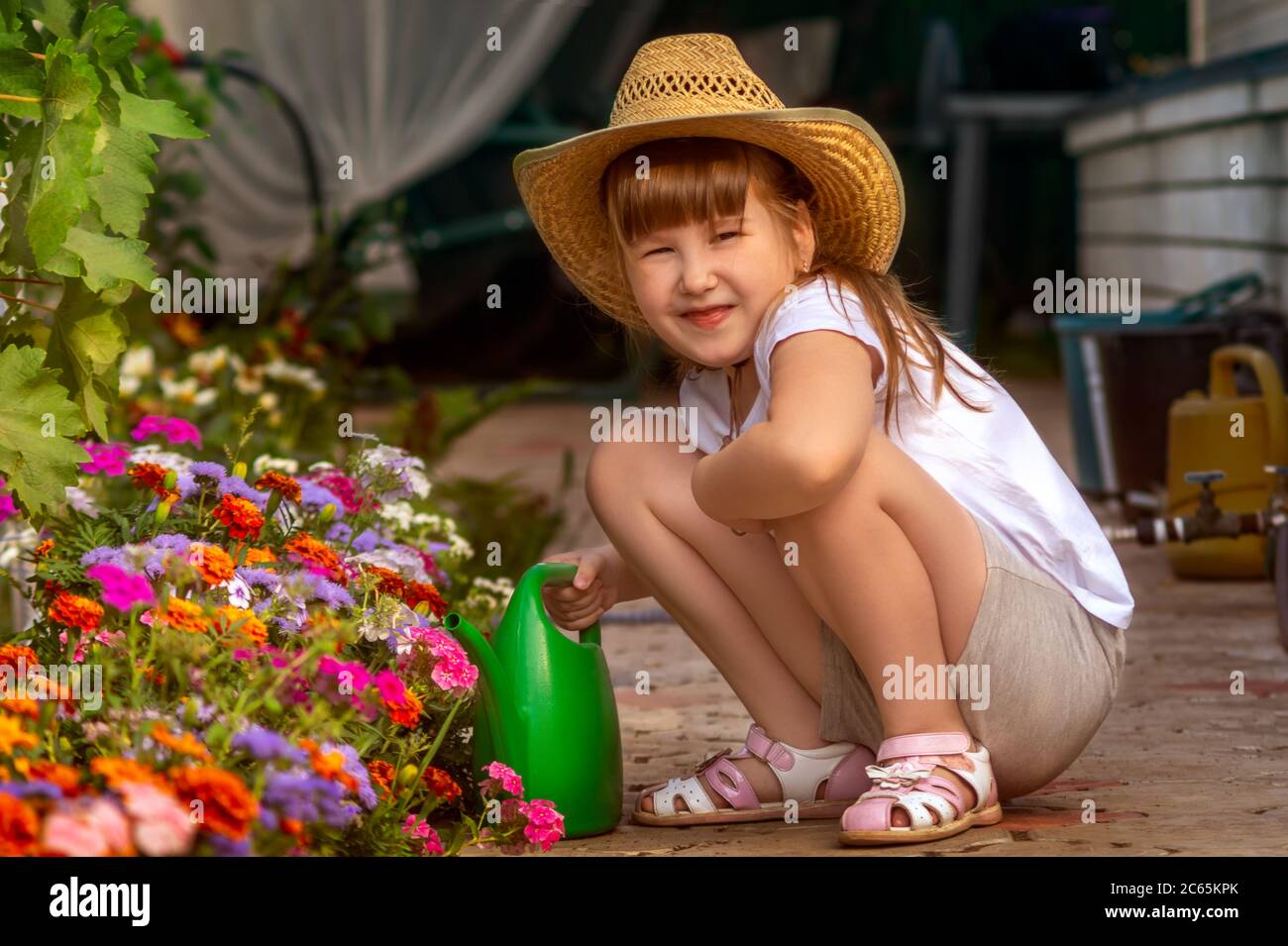 Jolie petite fille souriante en chapeau de cow-boy en paille en plein air dans le jardin prend soin du lit de fleur en l'arroser par le vert arroser peut. Banque D'Images