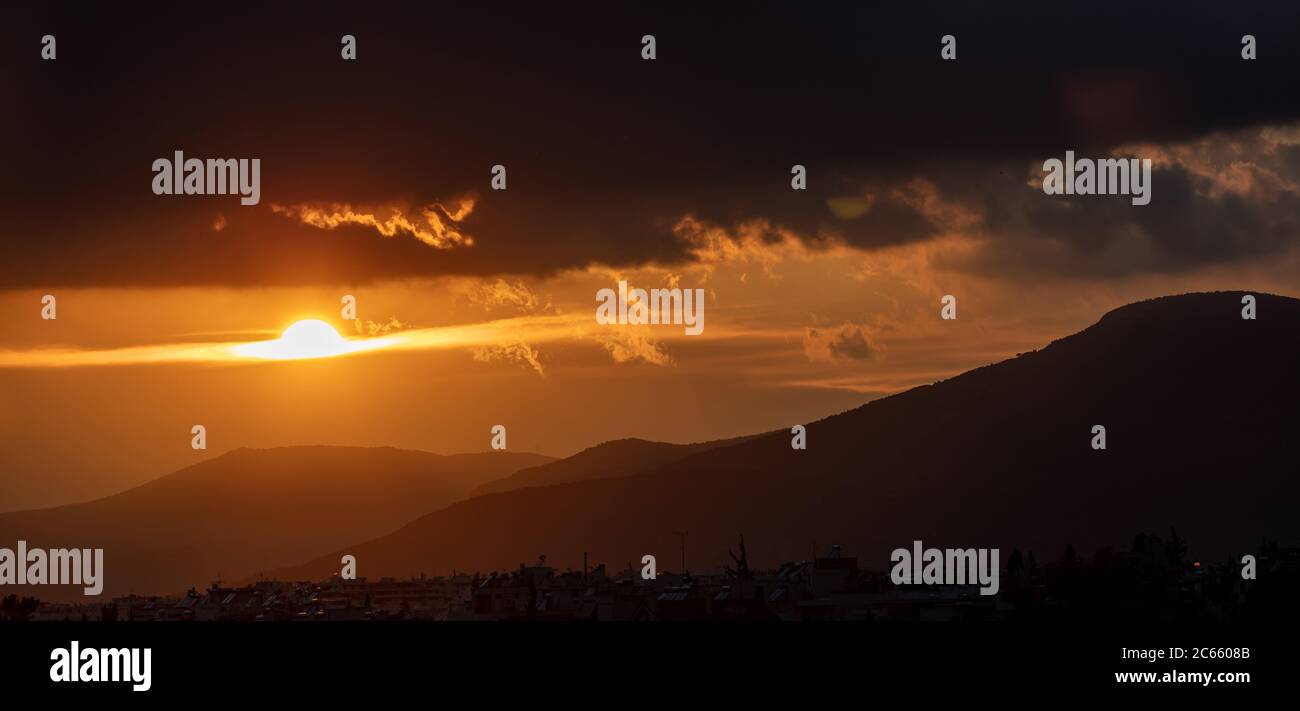 Coucher de soleil, lever du soleil à travers et derrière les nuages sur le fond des montagnes. Les poutres de soleil colorer le ciel les maisons du village et les collines silhouette noire. Banque D'Images