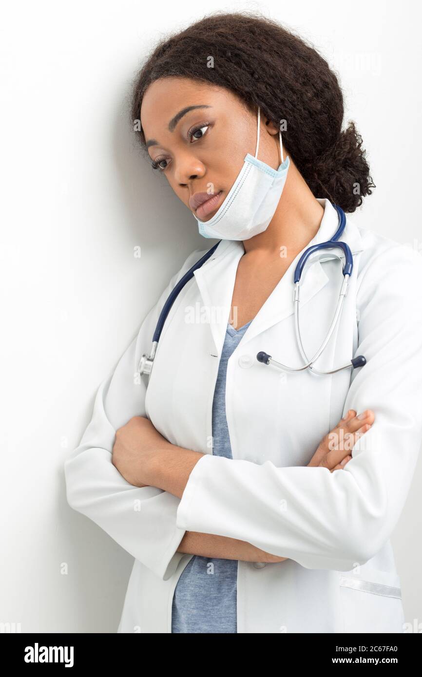 Portrait d'une femme afro-américaine fatiguée médecin en manteau blanc. Une infirmière triste dans un masque se tient près du mur de la clinique. Banque D'Images