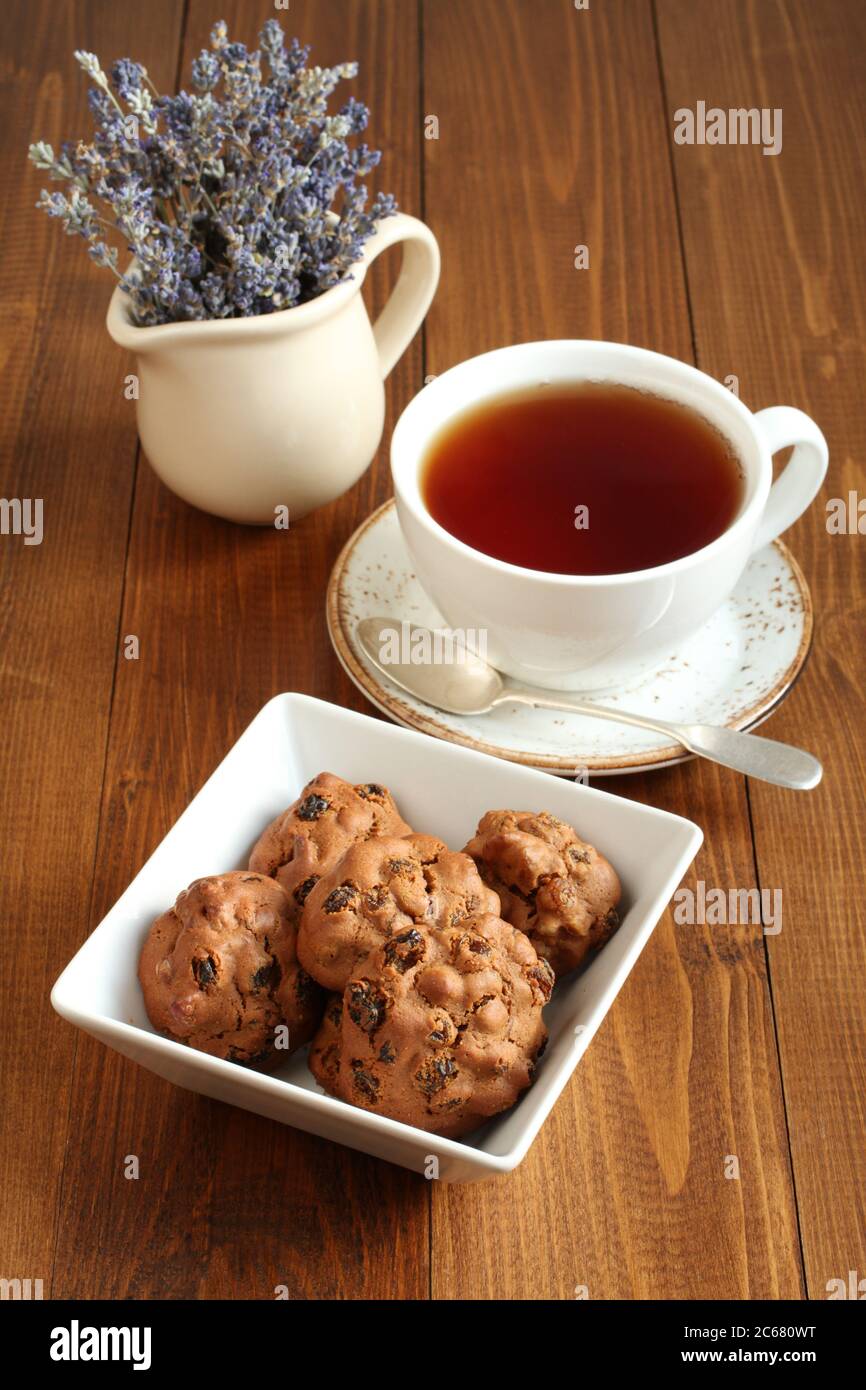 Biscuits au chocolat avec noix et raisins secs dans un bol carré et une tasse de thé noir sur une table en bois. Gros plan Banque D'Images