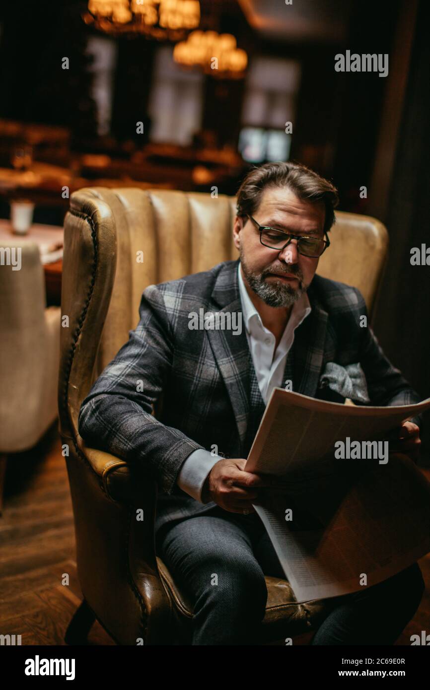 Homme adultes dans la cinquantaine, entrepreneur en costume et lunettes lisant le journal tout en restant assis dans le luxe véritable cuir fauteuil en r Banque D'Images