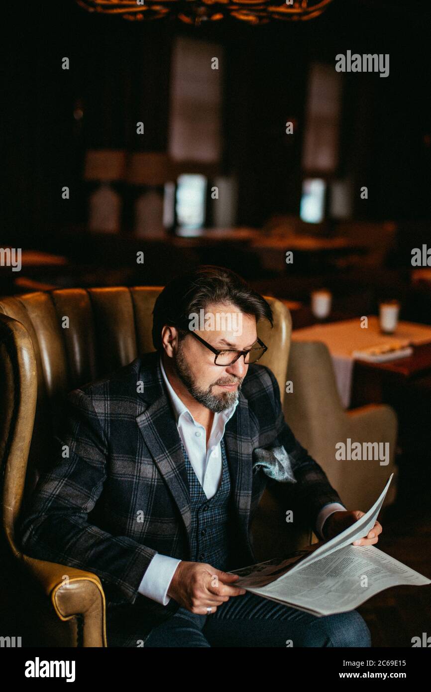 Pensive homme d'âge moyen dans un élégant costume sur mesure assis dans un fauteuil en cuir, la lecture de journaux frais dans la salle de séjour avec l'intérieur de luxe. Banque D'Images