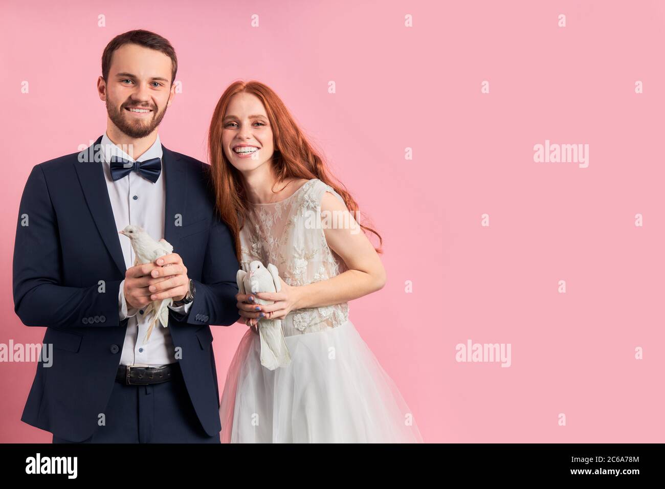 Belle femme avec les cheveux rouges et homme portant tuxedo tenant des pigeons dans les mains, heureux ensemble, stand isolé sur fond rose . Symbole colombe de h Banque D'Images