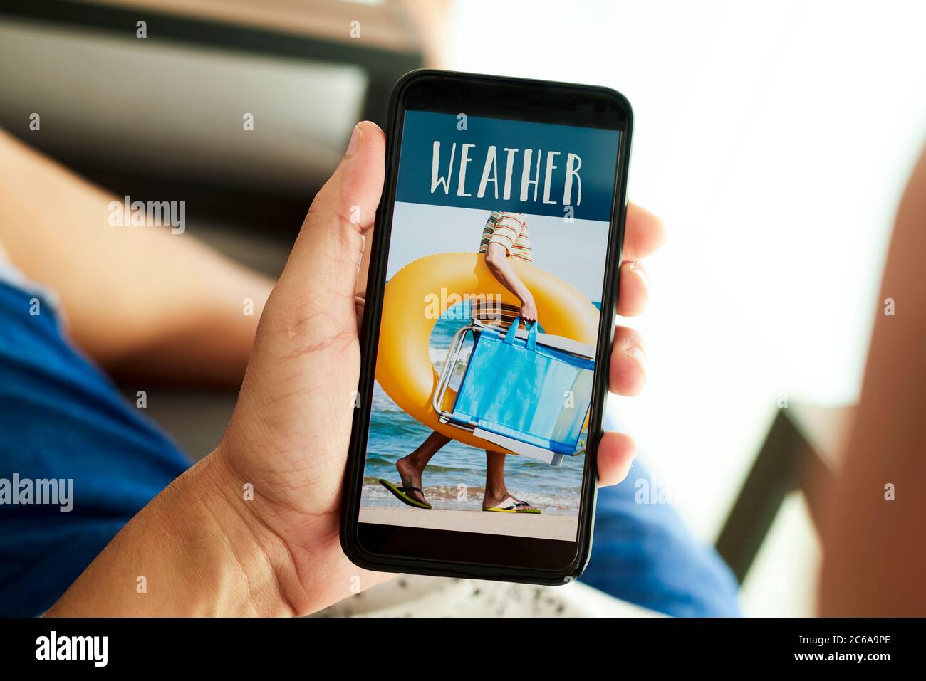 gros plan d'un jeune homme, portant des vêtements d'été décontractés, assis dans un fauteuil, ayant son smartphone dans ses mains avec le texte météo dans son écran Banque D'Images
