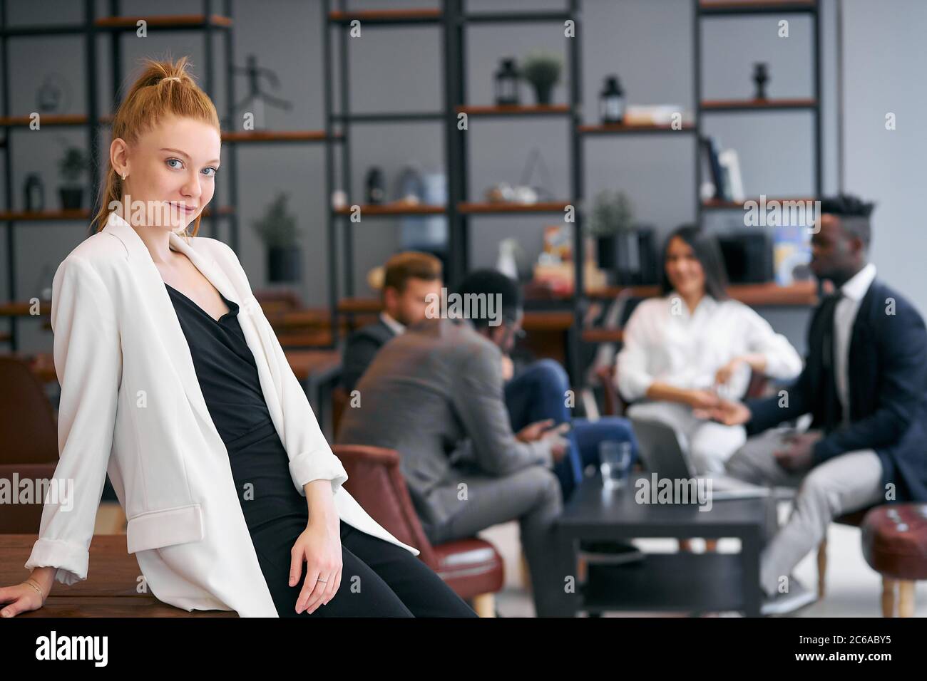 Une femme d'affaires souriante pose et regarde la caméra pendant que ses collègues ont une conversation derrière son arrière-plan moderne Banque D'Images