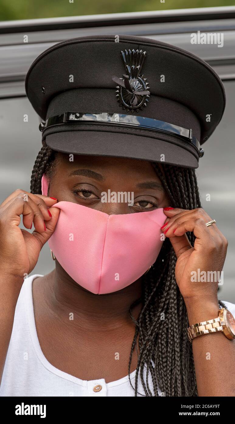Angleterre, Royaume-Uni. 2020. Portrait d'une femme chauffeur avec une casquette uniforme et des cheveux tressés ajustant son masque rose pendant l'épidémie de Covid-19 Banque D'Images