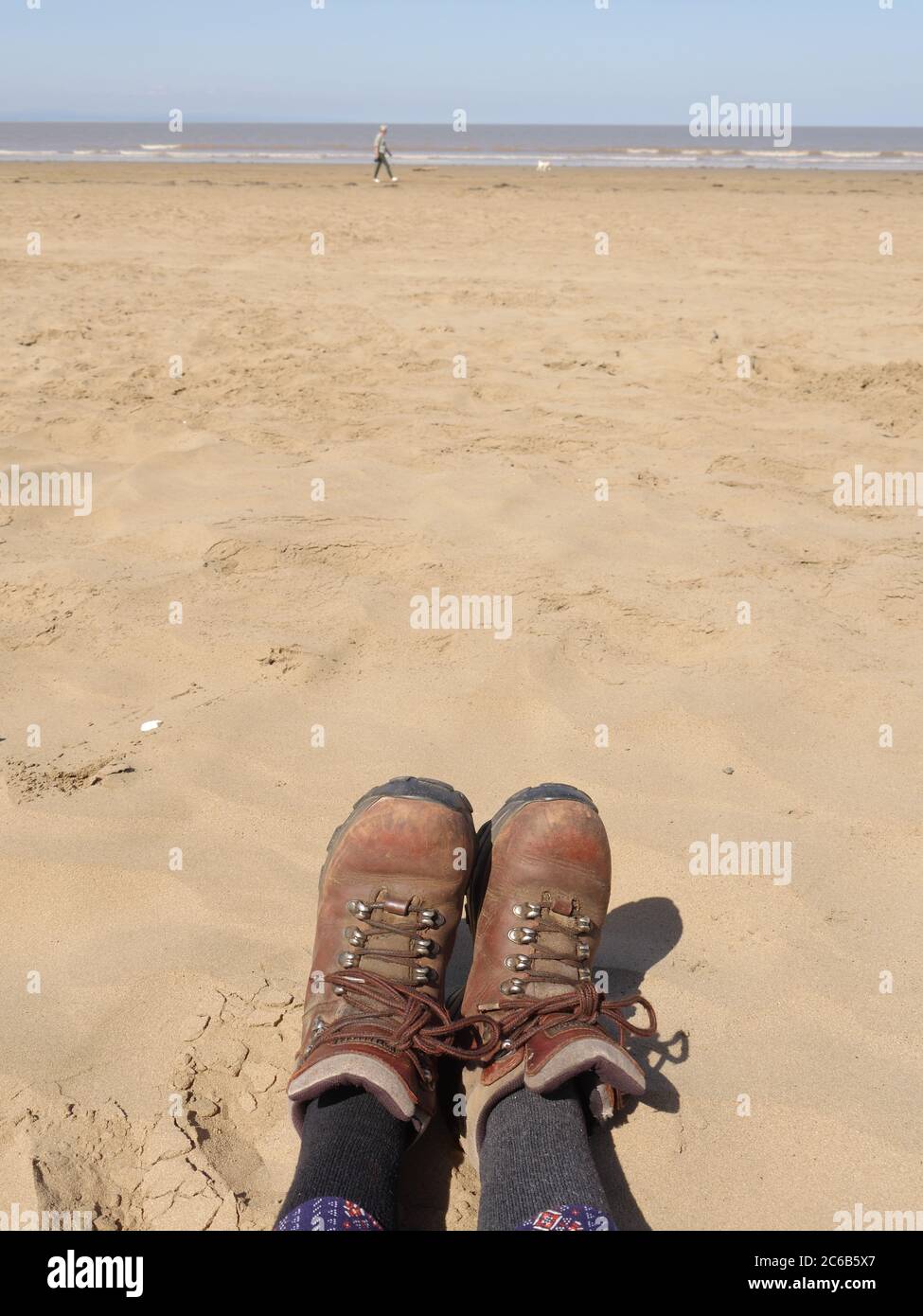 Vieilles bottes en cuir portées par une personne assise sur une plage de sable au loin une autre personne marchant un chien Banque D'Images