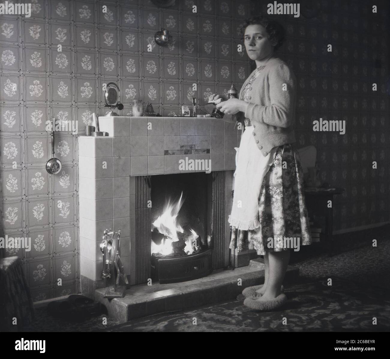 Années 1950, historique, une femme au foyer portant un tablier sur une robe à motifs floraux, un cardigan en laine et des pantoufles debout dans une pièce devant un feu de charbon ouvert dans une pièce de devant, nettoyant une horloge de la cheminée carrelée, Angleterre, Royaume-Uni. Banque D'Images