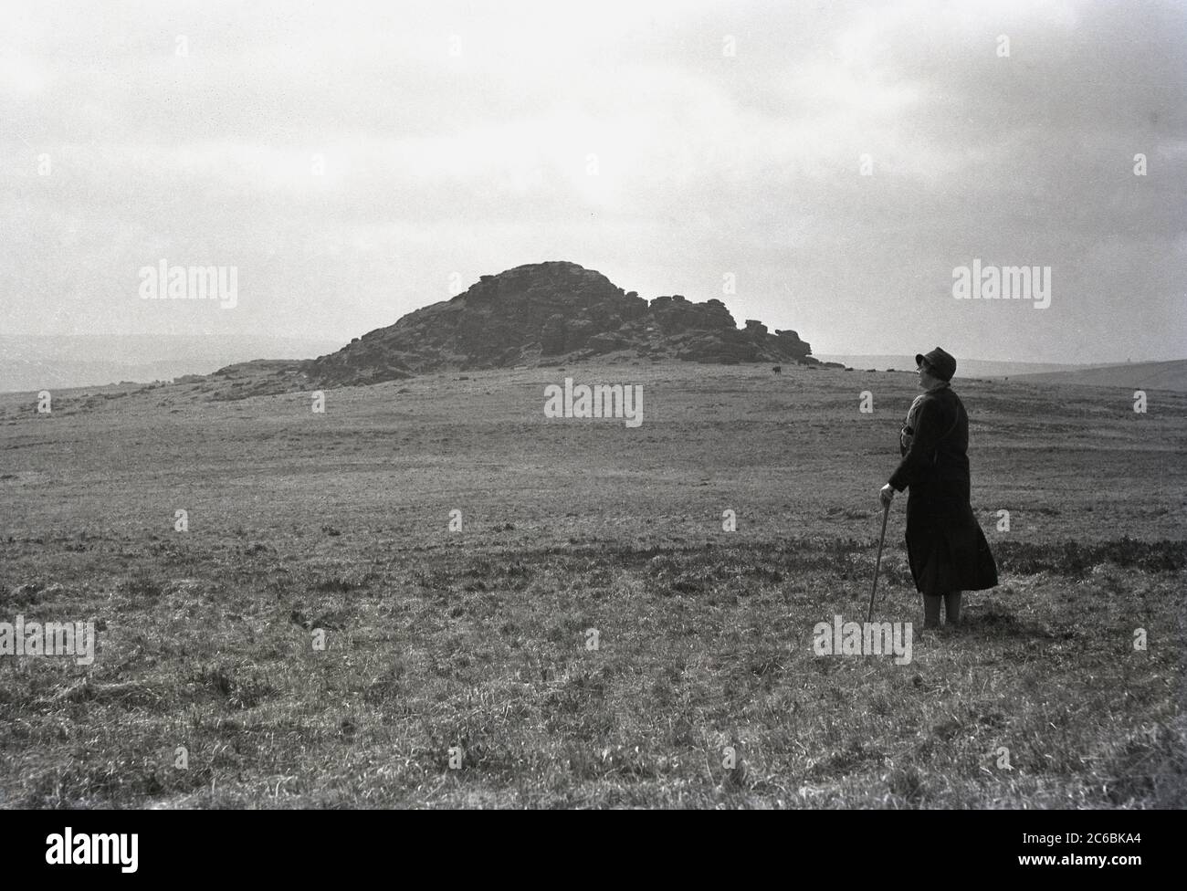 Années 1940, historique, une marcheuse mature, avec bâton, debout sur un sol plat regardant le paysage à Higher White Tor, Dartmoor, Devon, Angleterre, Royaume-Uni. L'un des tors supérieurs autour de la zone des deux ponts de la lande, situé sur une crête d'une colline signifie une vue magnifique. Les tors sont des affleurements de granit et le parc national de dartmoor en compte plus de 160. Banque D'Images