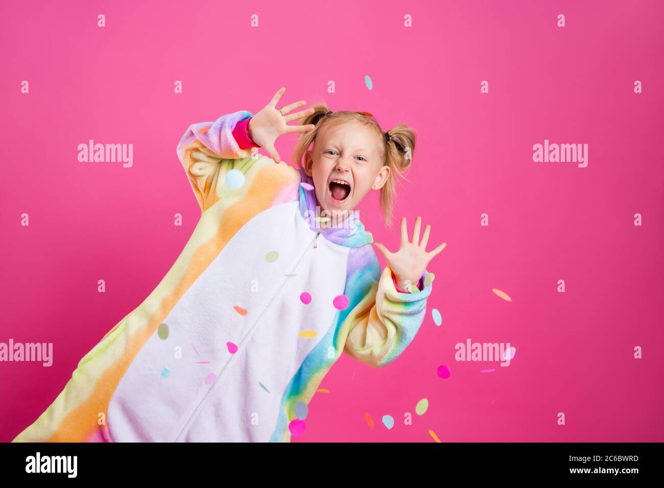 Bonne petite fille dans kigurumi unicorn sur fond rose se réjouit dans les confettis multicolores, espace pour le texte Banque D'Images