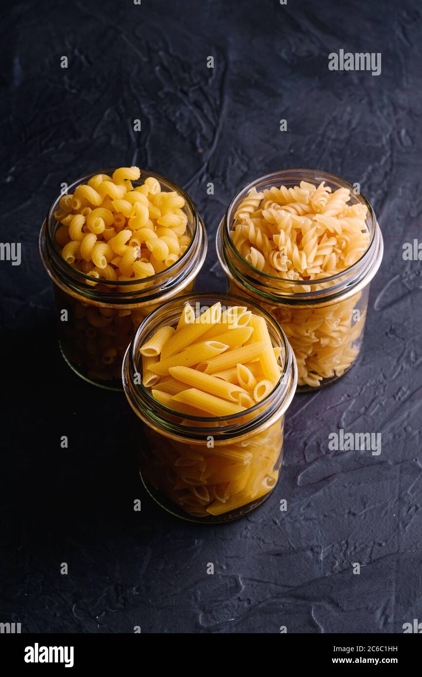 Trois pots en verre avec variété de pâtes de blé doré non cuites sur fond noir foncé texturé, vue en angle Banque D'Images