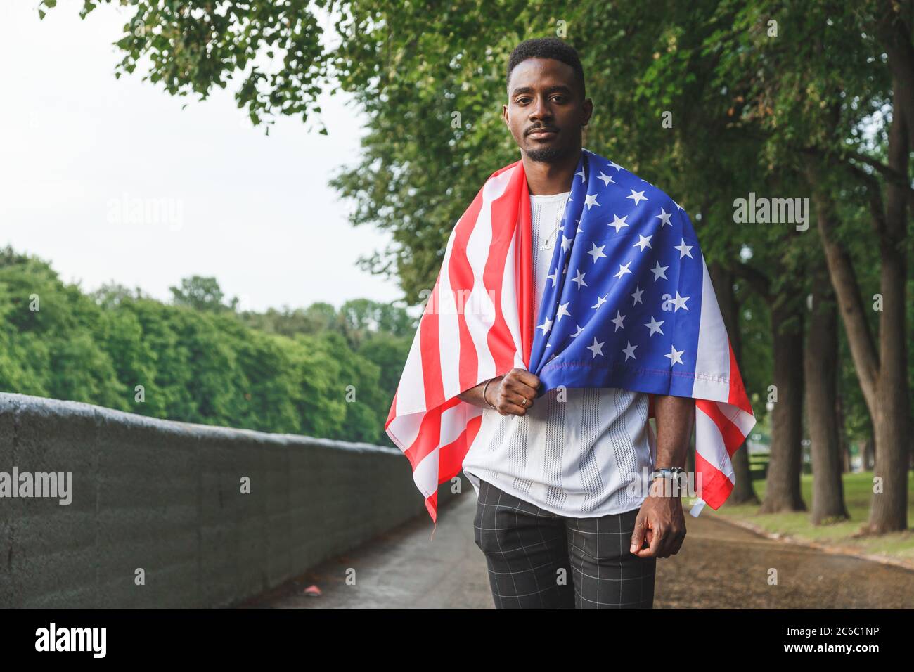 Beau Afro américain homme avec drapeau des Etats-Unis sur ses épaules regardant sérieusement la caméra, debout à l'extérieur. Jour d'été Banque D'Images