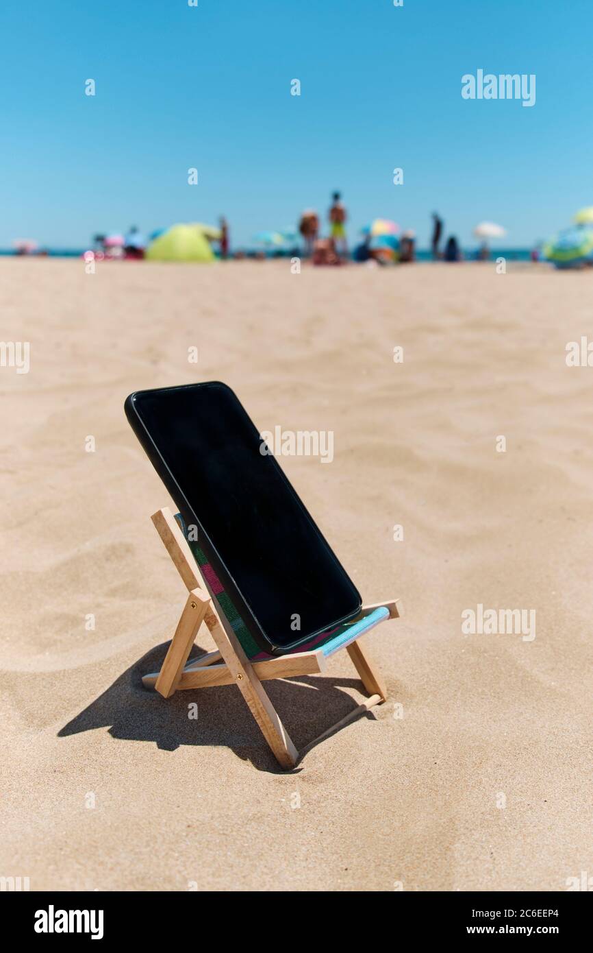 un smartphone noir dans une chaise longue sur le sable de la plage, représentant le concept numérique de détox, avec des personnes méconnaissables appréciant et la mer en t Banque D'Images