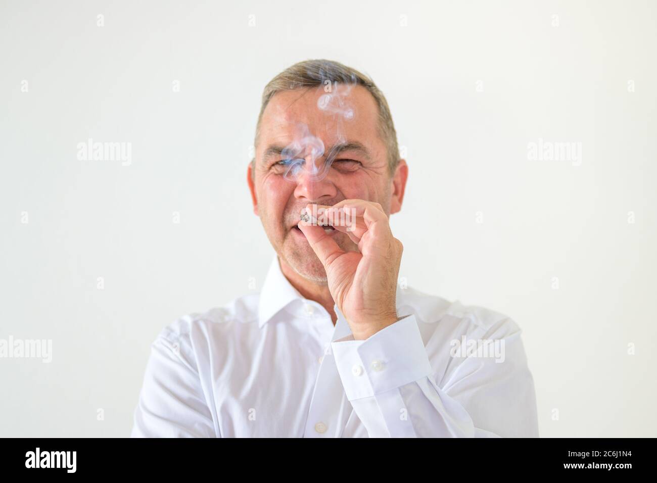 Homme réfléchi qui se paffent sur une cigarette qui se concentre derrière la fumée dans un portrait de tête et d'épaules sur blanc Banque D'Images