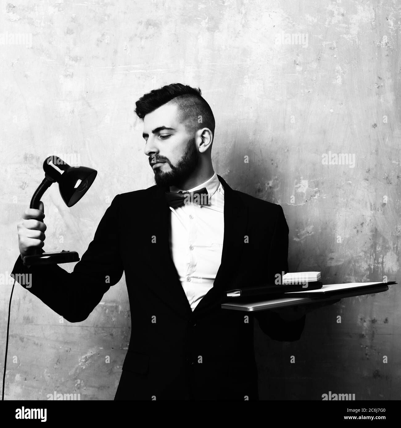 Broker avec une barbe soignée, style et expression du visage satisfait regarde la lampe de bureau et tient l'ordinateur portable et la papeterie à portée de main, sur le vieux fond beige mur. Concept de travail au bureau Banque D'Images