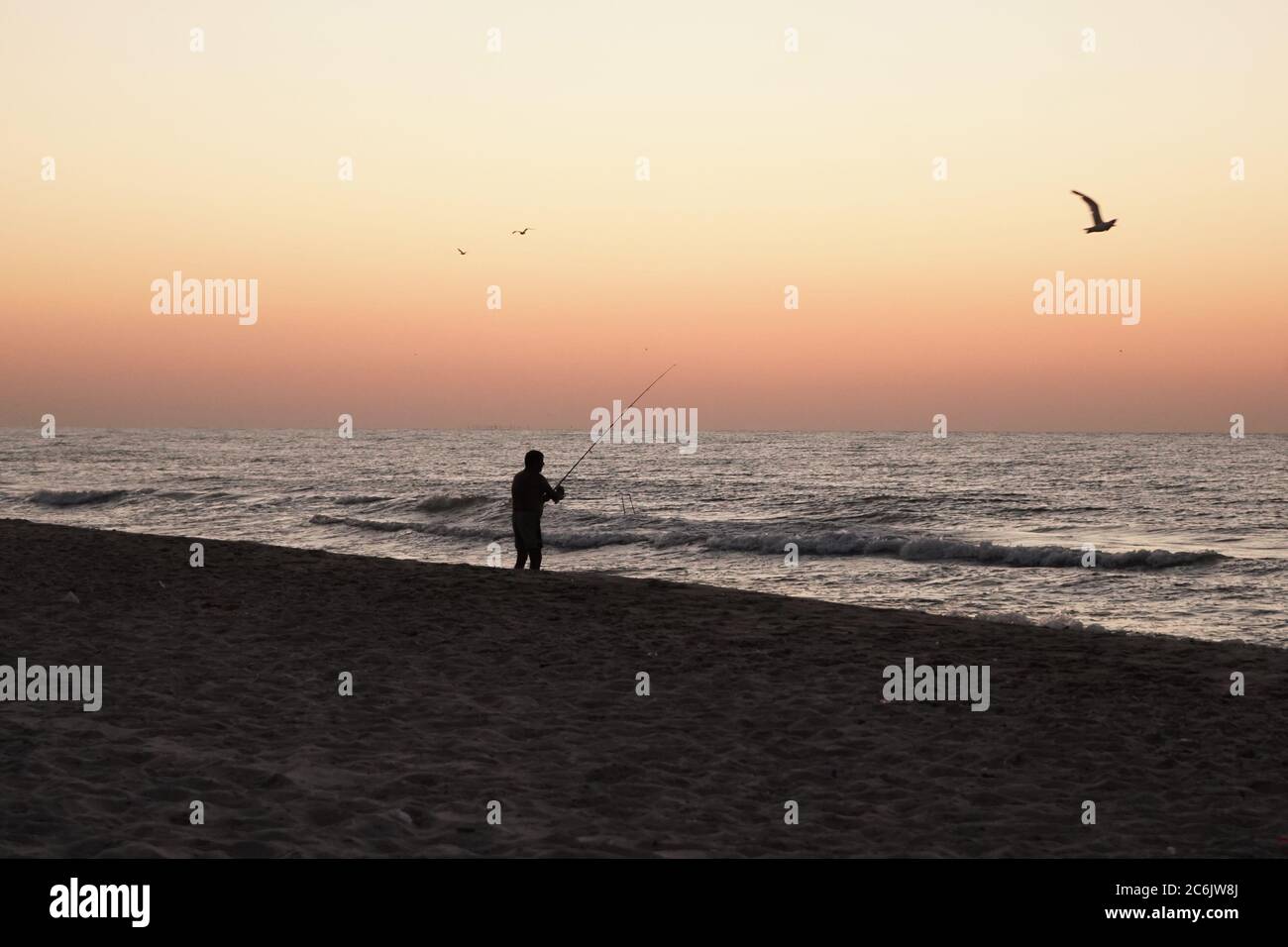 Le pêcheur capture du poisson depuis la rive au coucher du soleil. Le vieil homme tient la canne à pêche dans ses mains tout en se tenant sur la côte de la mer. Silhouette masculine sombre près Banque D'Images