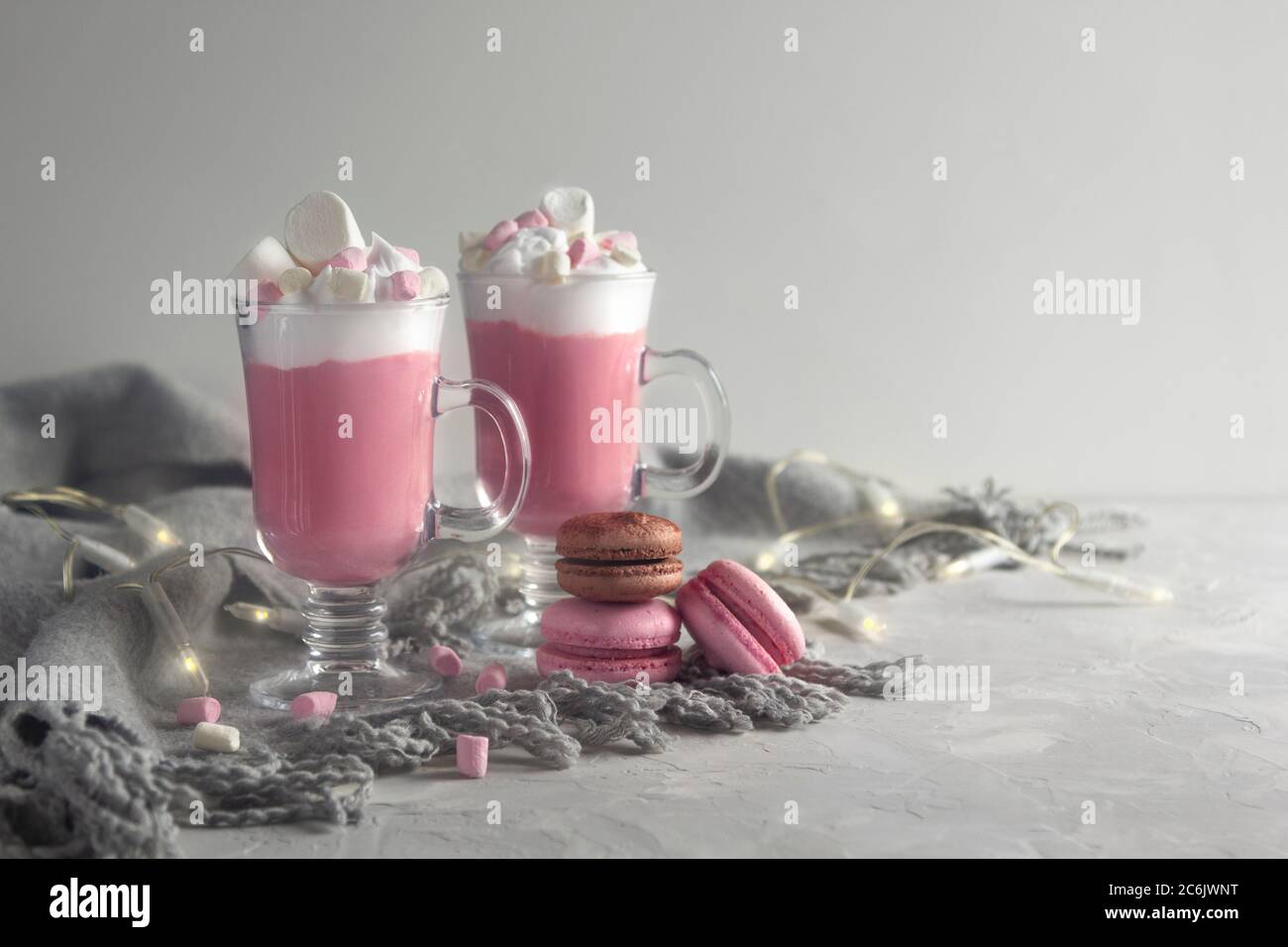 Chocolat chaud rose avec crème fouettée et bonbons guimauve dans 2 tasses en verre, macarons français doux pastel et lumières. Copier l'espace pour le texte. Horizont Banque D'Images