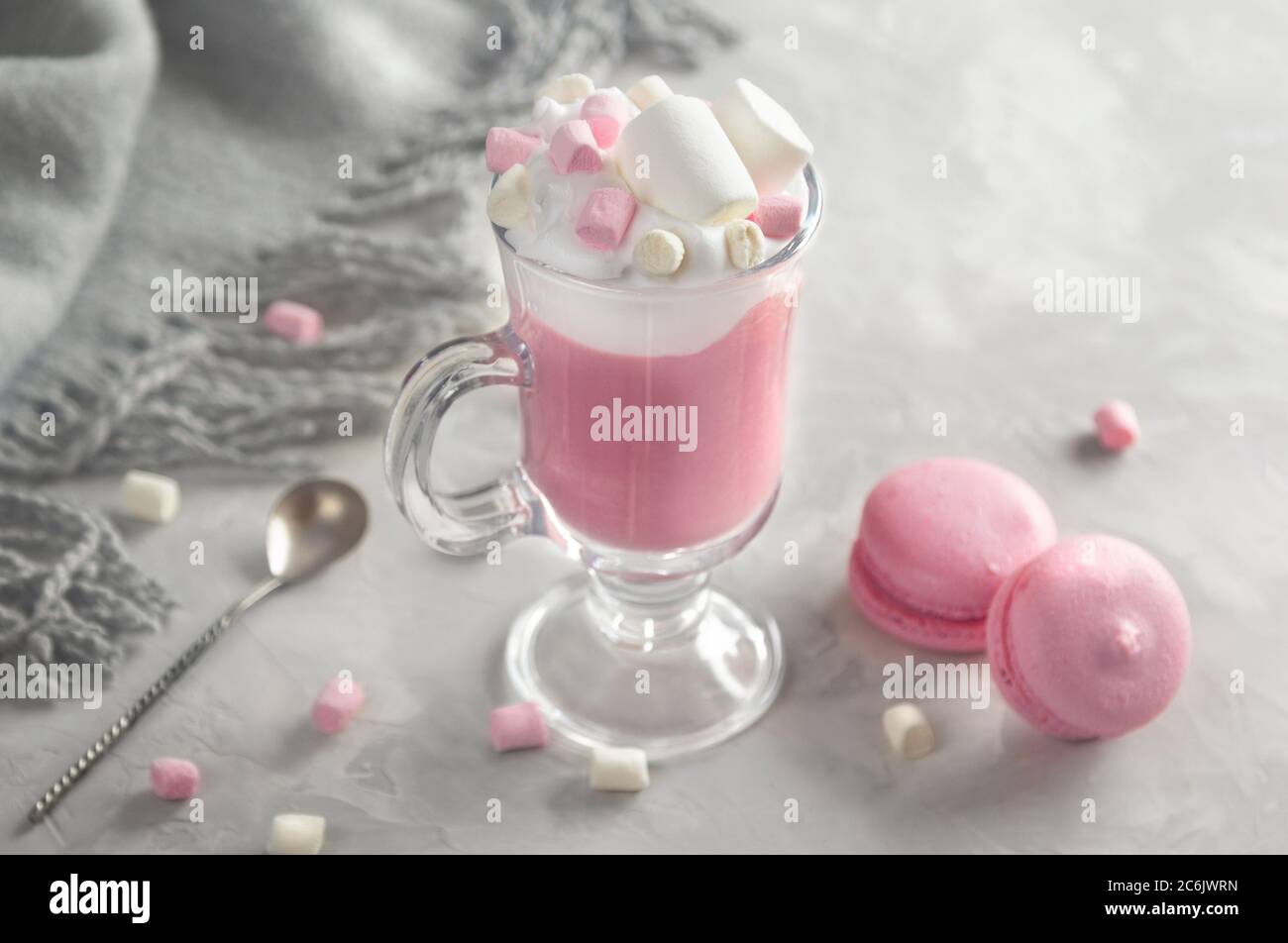 Chocolat chaud rose avec crème fouettée et bonbons guimauve dans une tasse de verre, macarons français doux pastel sur fond gris. Banque D'Images