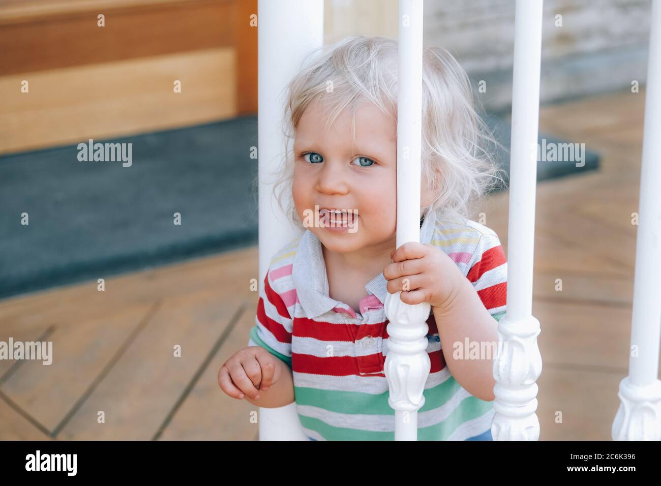 Portrait d'un joli petit garçon heureux avec des cheveux blonds et des yeux bleus, debout derrière les rails d'escalier blancs dans le café extérieur, se laquant comment marcher. Retour au concept normal. Banque D'Images