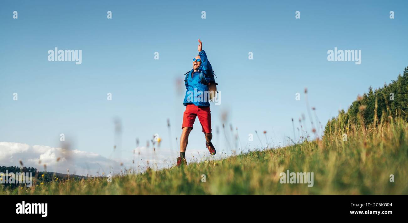 Le voyageur de Backpacker sautant émotionnellement au-dessus de la prairie de montagne d'herbe verte avec un sac à dos avec de larges bras et jambes ouverts Banque D'Images