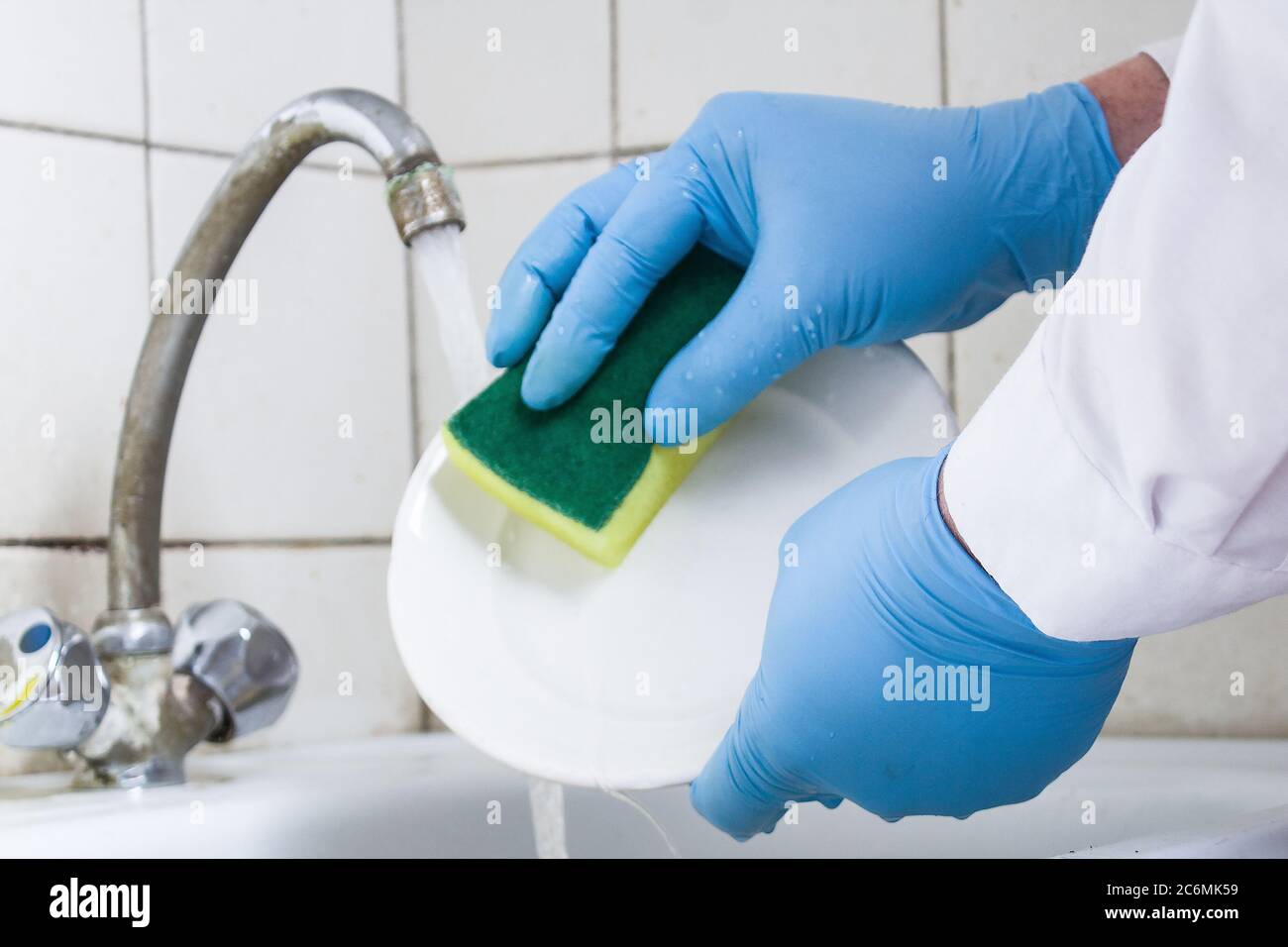 gants bleus mains laver une assiette blanche sur l'évier de cuisine contre un mur de carrelage blanc Banque D'Images