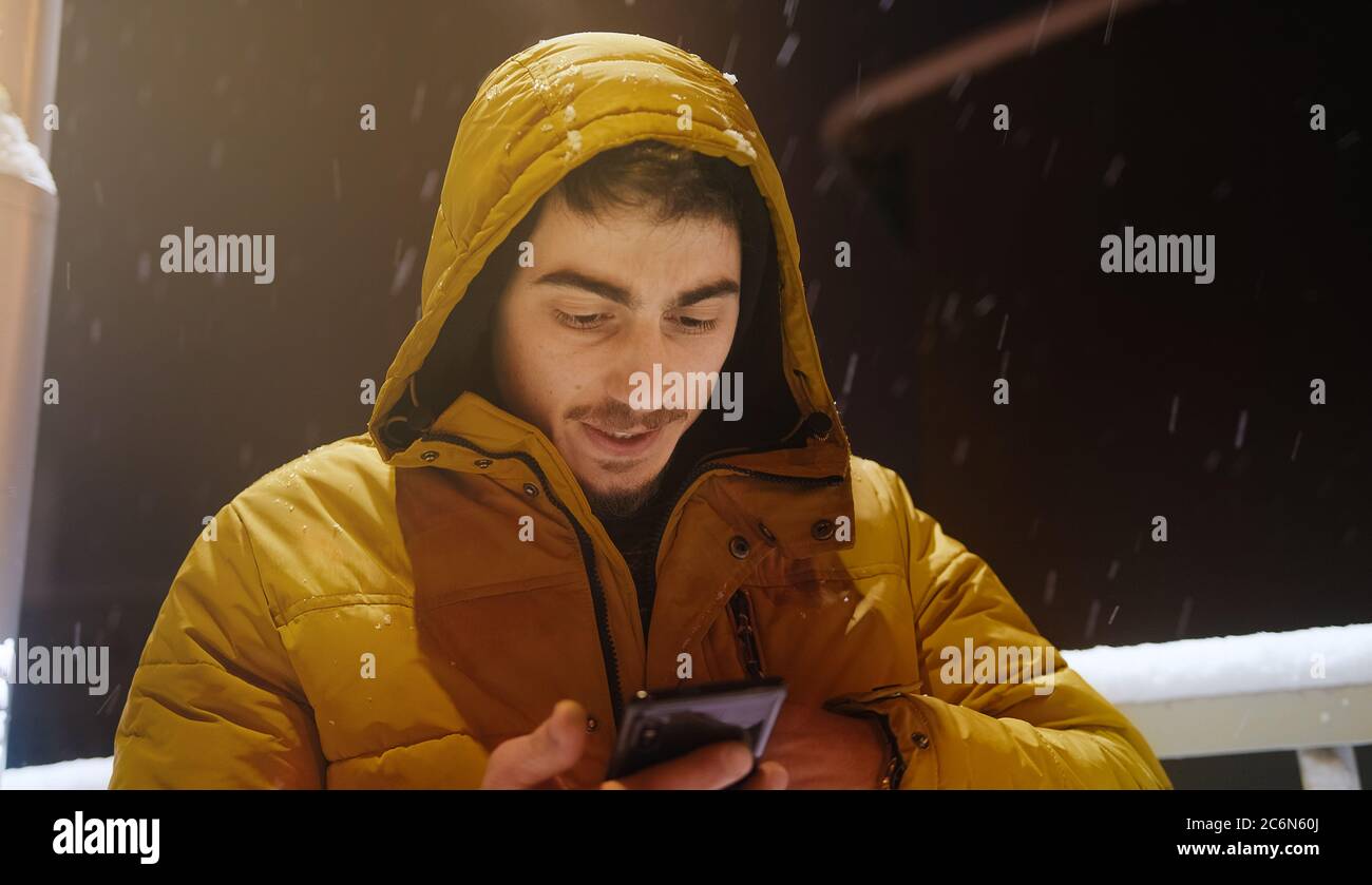 Un jeune homme à capuche parle dans la rue par téléphone pendant qu'il neige en hiver Banque D'Images