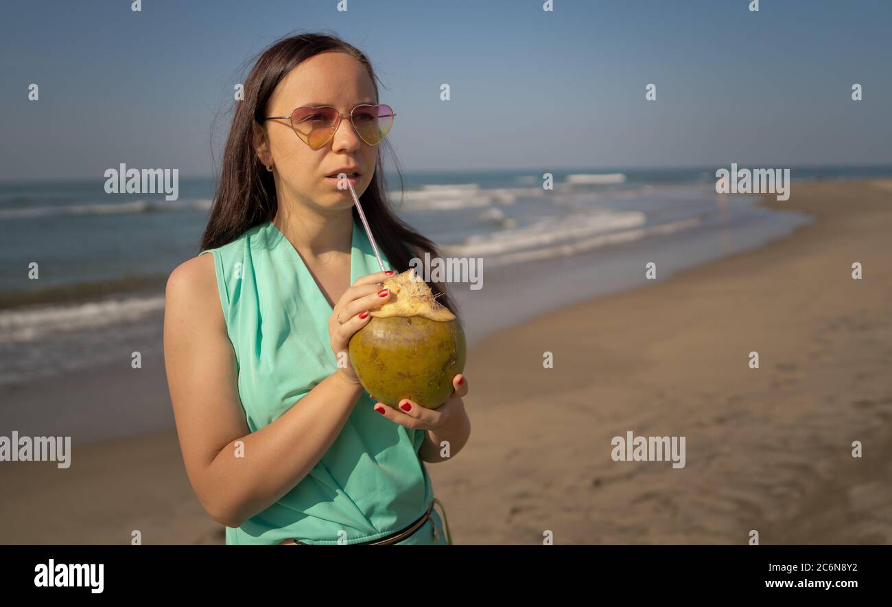 Jeune femme en lunettes de soleil qui boit de la noix de coco près de la mer ou de l'océan. Femme touriste appréciant une boisson tropicale, sirotant de l'eau de noix de coco à travers la paille sur le sable Banque D'Images