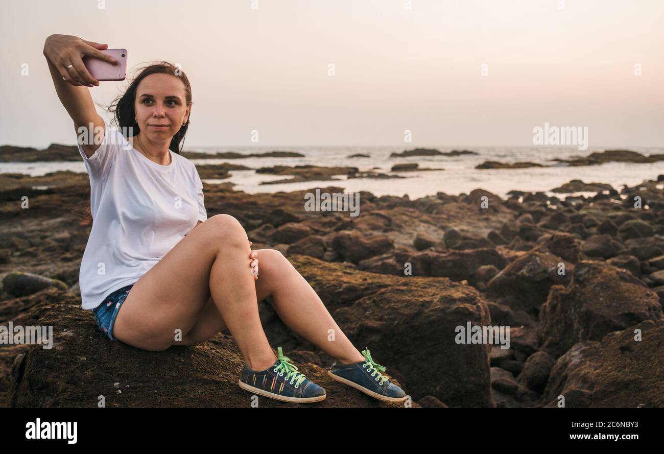 Une belle jeune femme prend le selfie sur un téléphone portable, assis sur les rochers au bord de la mer contre le coucher du soleil. Banque D'Images