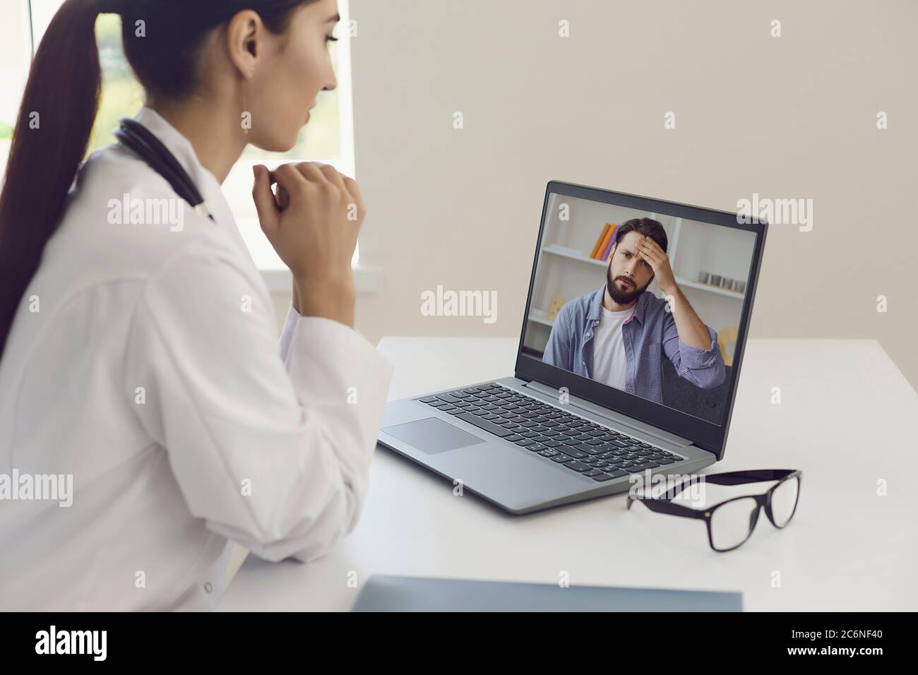 Médecin en ligne. Médecin à l'écoute d'un patient souffrant d'un mal de tête d'un groupe de sympomes utilise un ordinateur portable de chat vidéo sur une table dans un bureau de clinique. Banque D'Images
