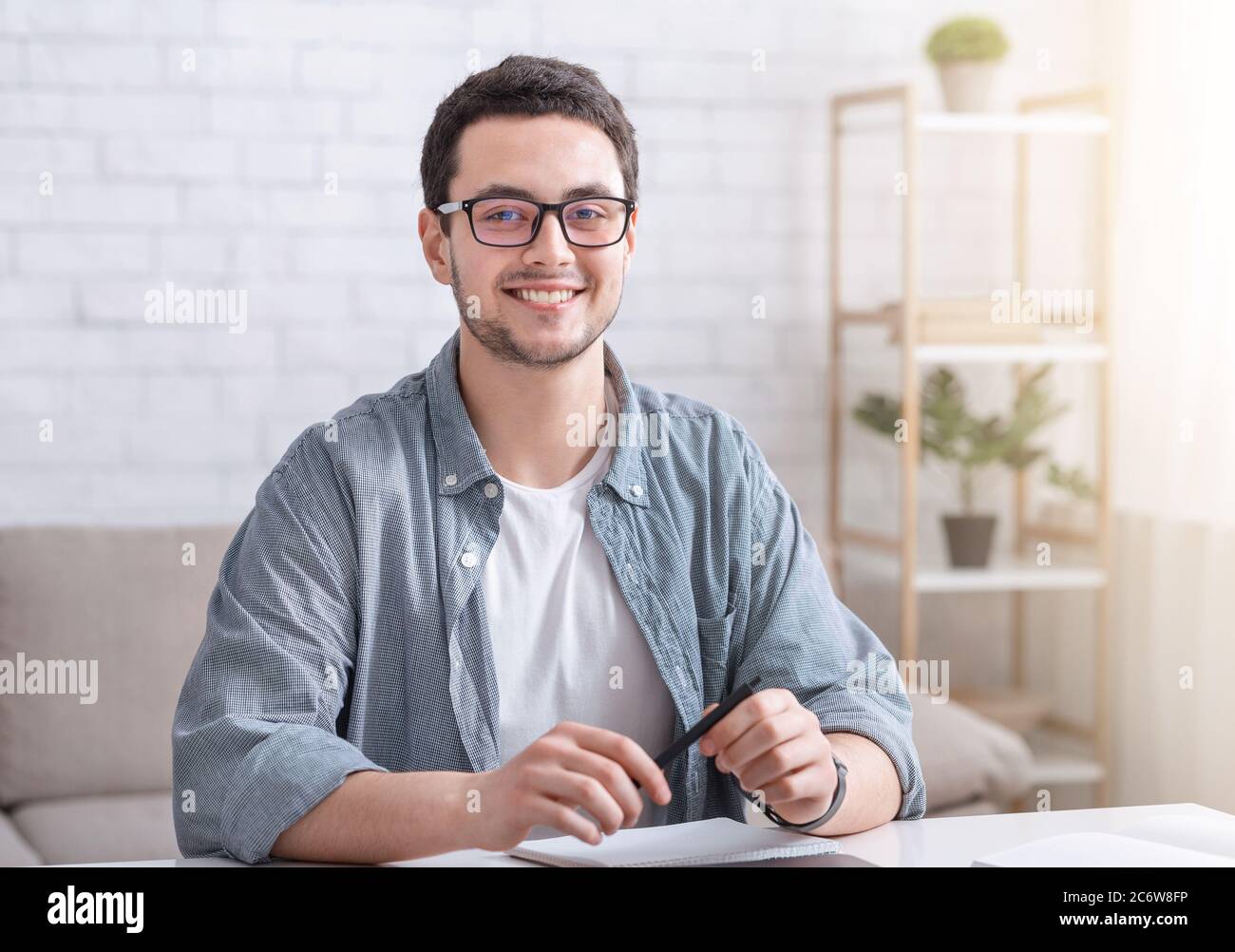 Étude en ligne à domicile pour les étudiants. Un homme souriant avec des lunettes est assis à la table dans le salon intérieur Banque D'Images