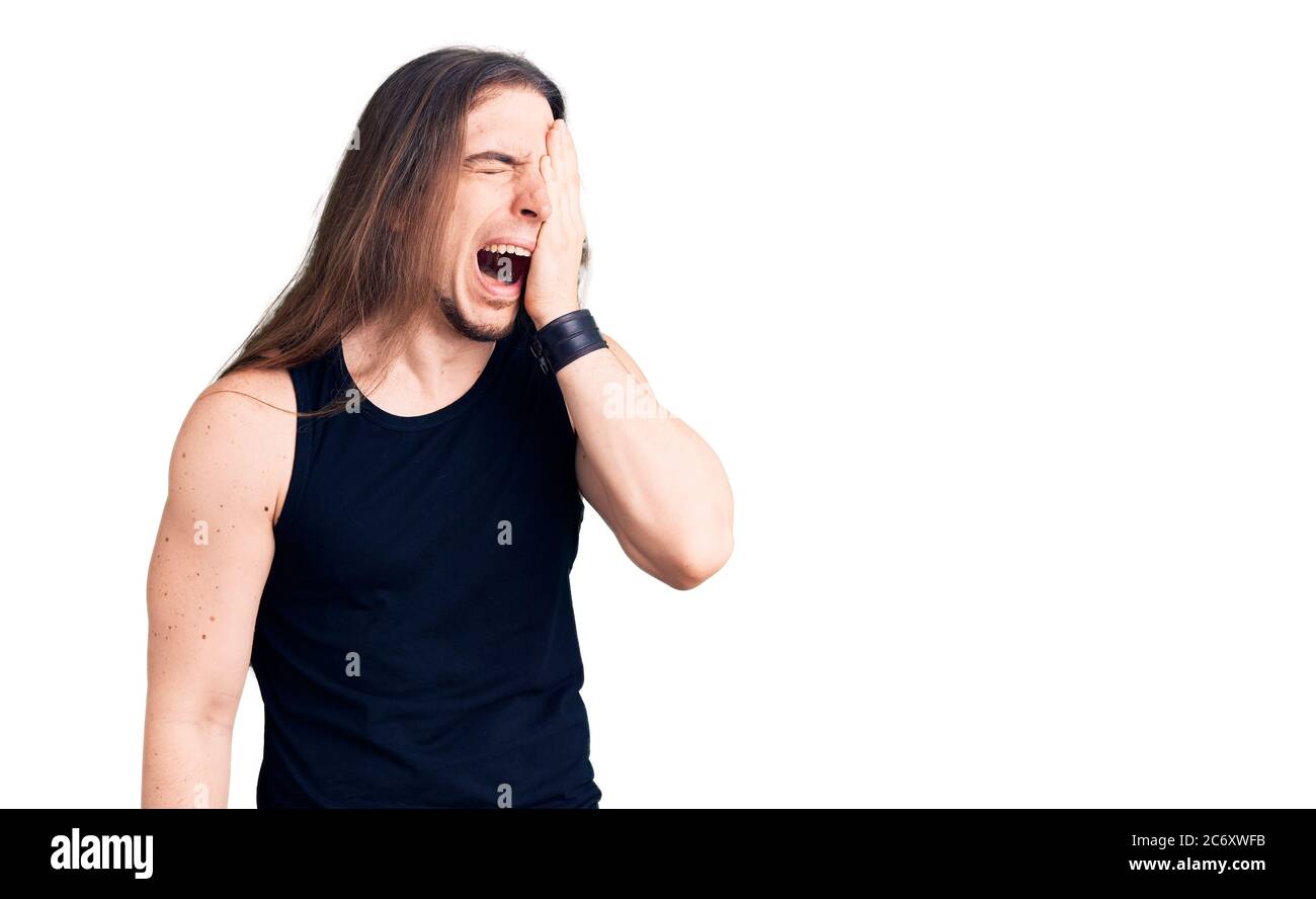 Jeune homme adulte avec de longs cheveux portant le style goth avec des vêtements noirs bâilling fatigué couvrant la moitié du visage, des yeux et de la bouche avec la main. Le visage fait mal dans la douleur. Banque D'Images