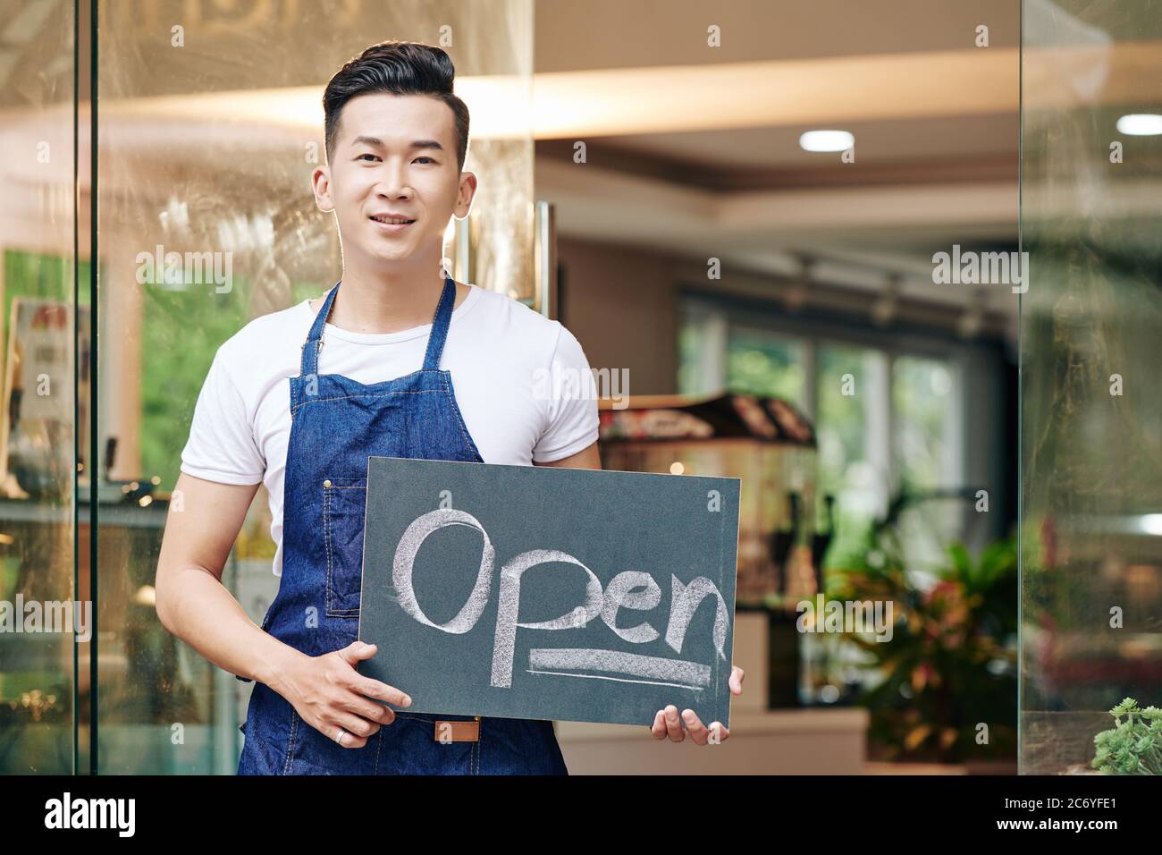 Jeune homme asiatique positif debout à l'entrée du café avec panneau ouvert Banque D'Images
