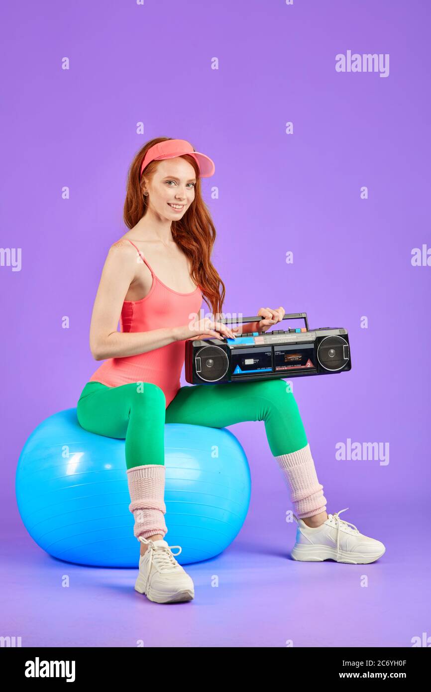 femme posant pour photoshoot style rétro, assise sur le ballon de fitness bleu avec lecteur de cassette portable sur ses genoux, vêtu d'un confortable bo rose Banque D'Images