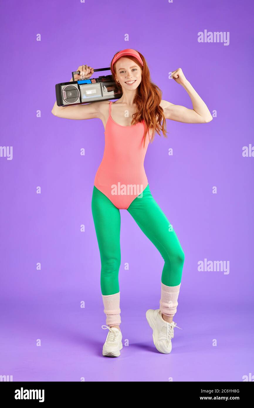 photo pleine longueur d'une jeune fille au gingembre, qui se tient debout avec un lecteur audio portable sur son épaule et qui montre ses biceps, en étant fière de soi et souriante happ Banque D'Images