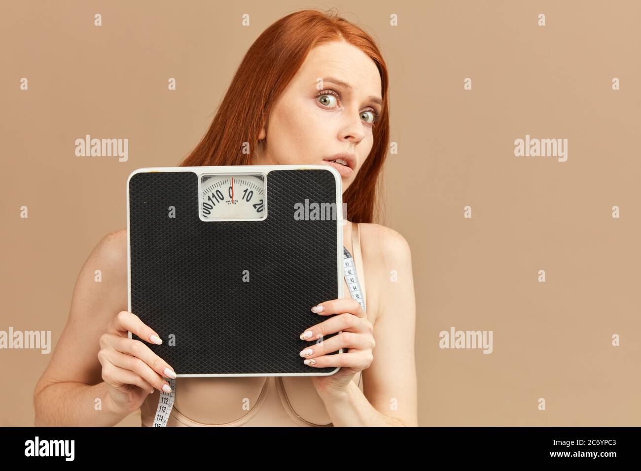 Timide effrayé rouillé femme skinny à cheveux rouges avec les yeux bougés, se cachant face derrière les balances mécaniques, isolé sur fond beige Banque D'Images