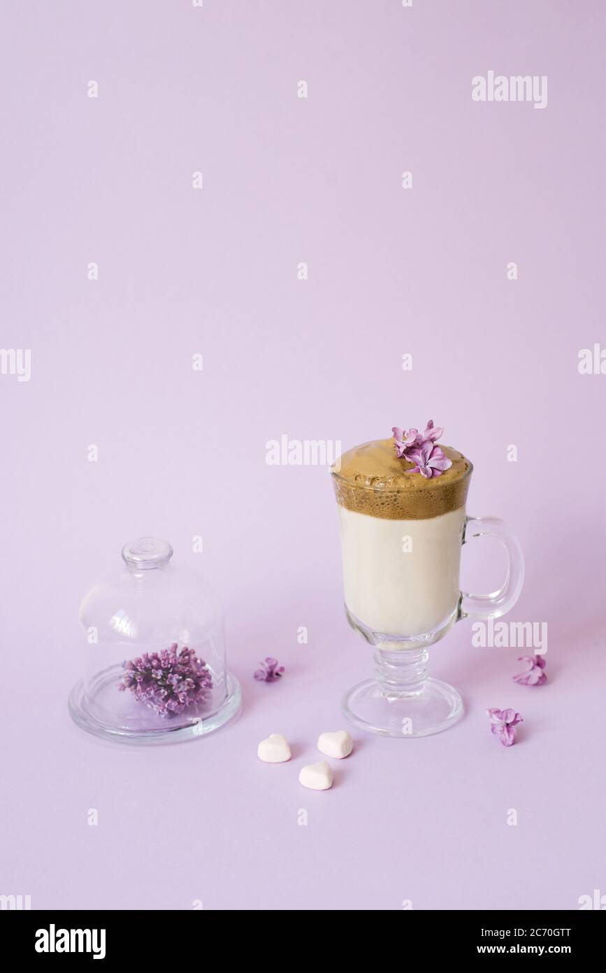 Dalgona café dans une tasse transparente avec tube en papier, une assiette de fleurs lilas et de bonbons sur fond lilas avec un espace pour copier Banque D'Images