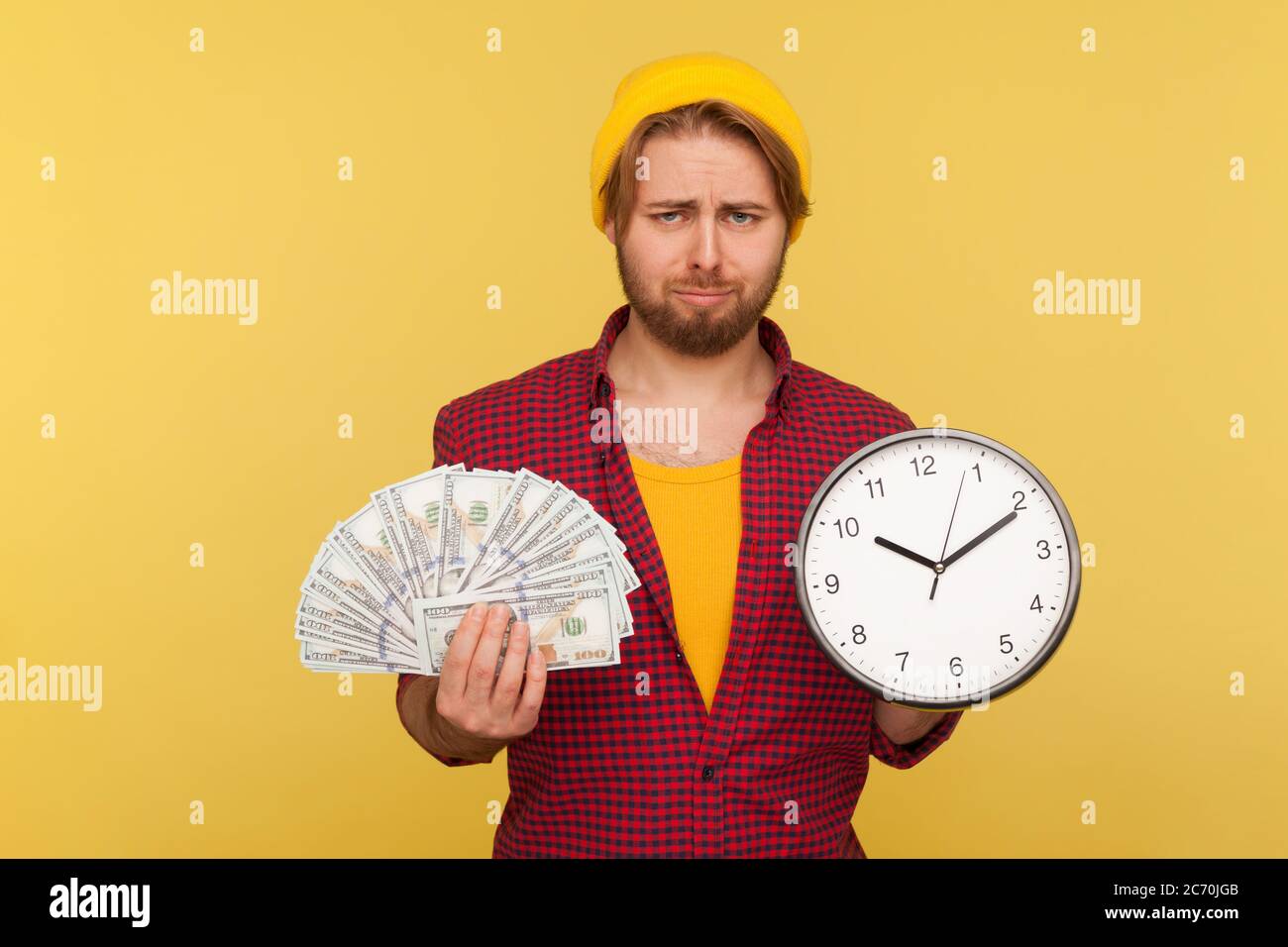 Contrarié hipster barbu gars dans la chemise à carreaux tenant une grande horloge et des billets de dollars, regardant la caméra avec une expression frustrée malheureux, est au sujet de t Banque D'Images