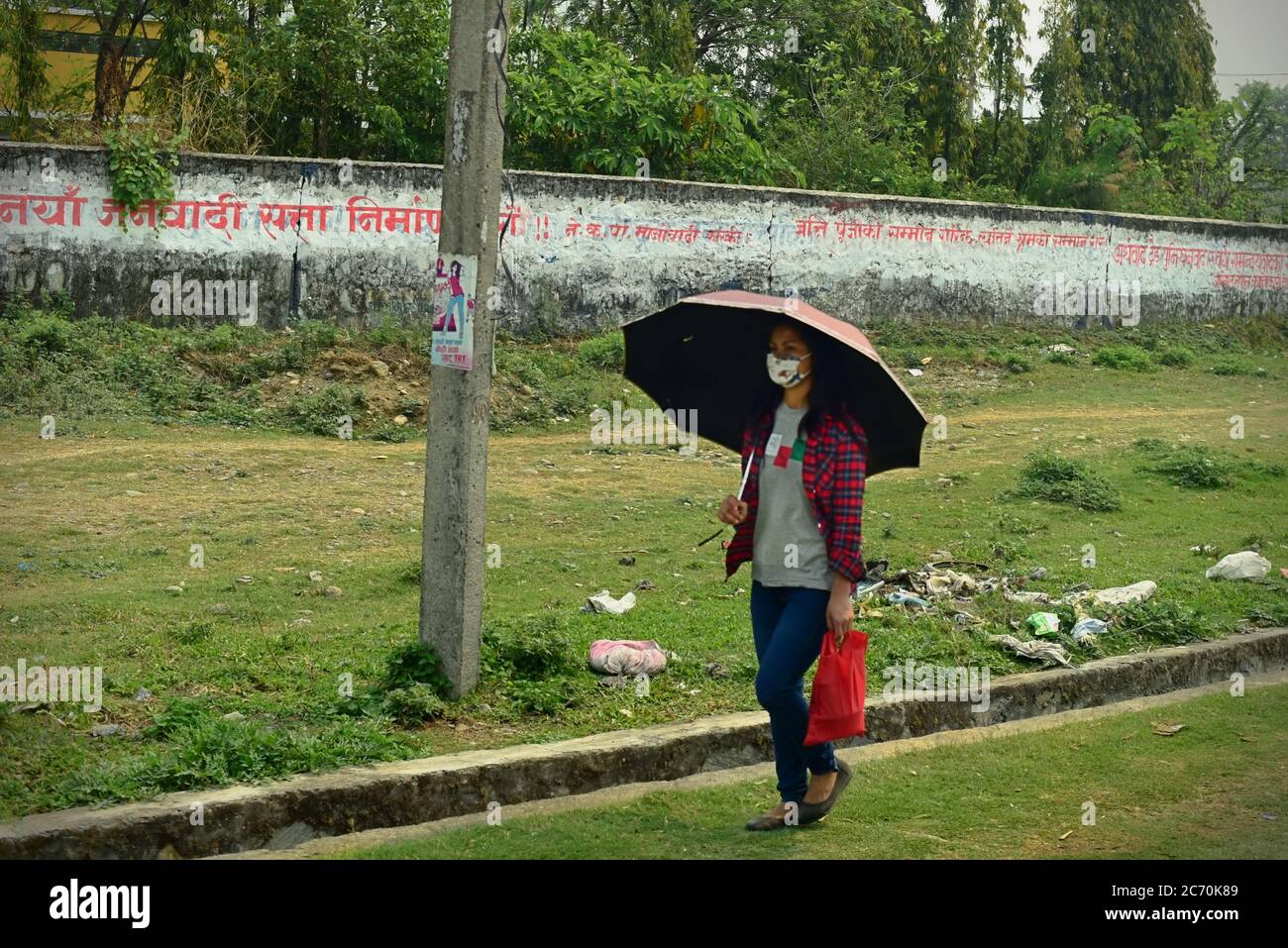 Une femme portant un masque facial, portant un parapluie alors qu'elle marche sur un bord de route dans la province de Bagmati Pradesh, au Népal. Banque D'Images