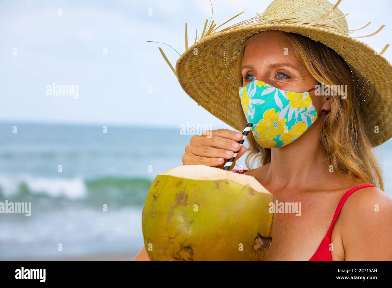 Portrait drôle de femme en chapeau de paille buvant une jeune noix de coco sur la plage tropicale de mer. Règles pour porter un masque facial dans les lieux publics en raison du coronavirus covid 19 Banque D'Images