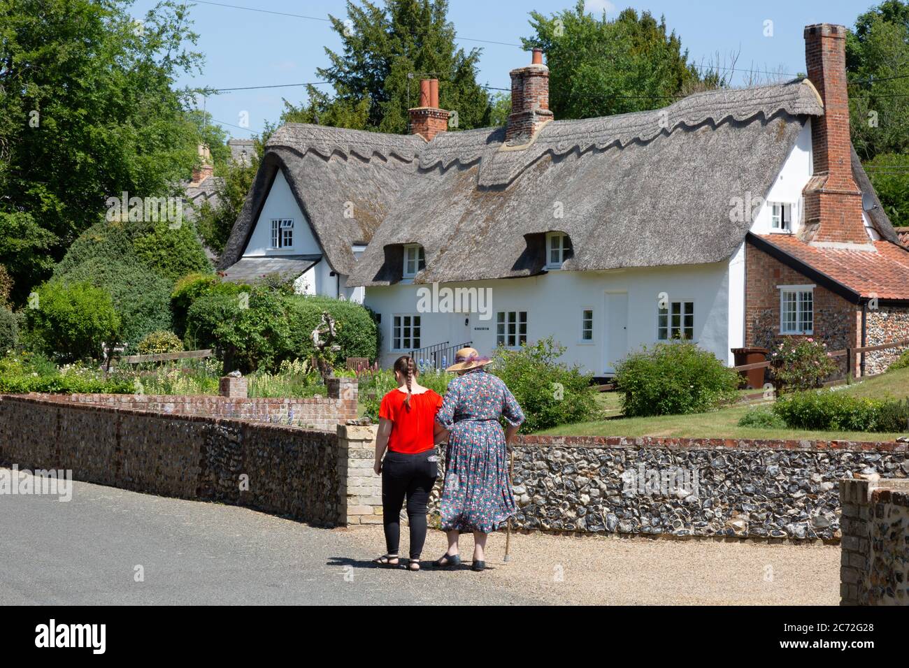 Scène de rue de village anglais en été; deux femmes marchant dans une rue devant un cottage traditionnel de chaume, village de Dalham, Suffolk Angleterre Royaume-Uni Banque D'Images