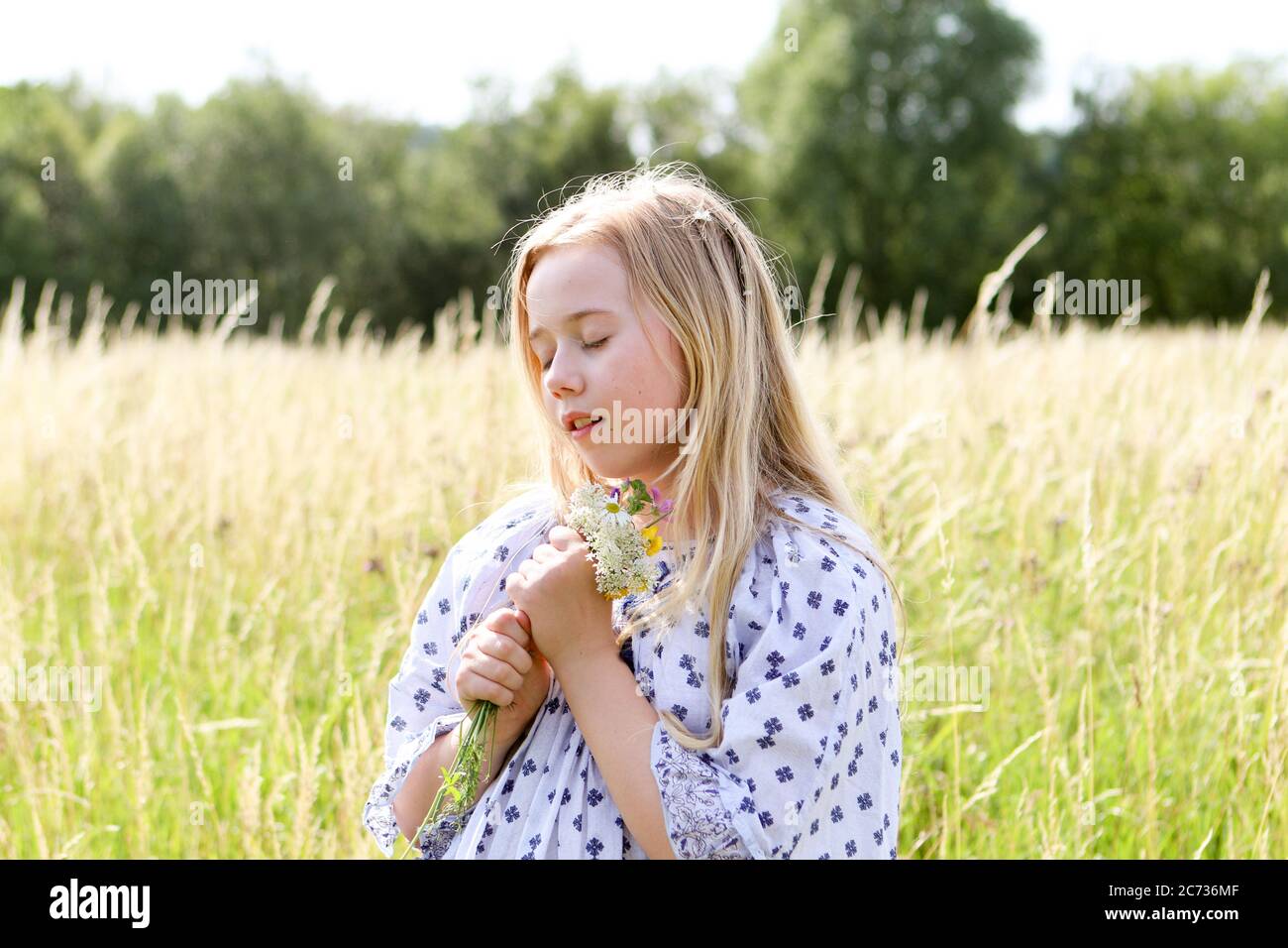 Une jeune fille assez hippie avec une guirlande dans ses cheveux blonds tient une posy de fleurs sauvages au soleil dans un champ de pâturage en été. Banque D'Images