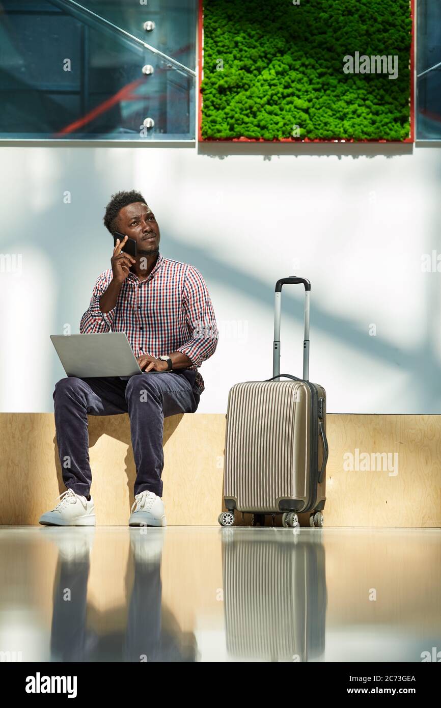Un jeune homme d'affaires africain parlant sur un téléphone portable et un ordinateur portable pendant qu'il est assis à l'aéroport il commandant des billets d'avion Banque D'Images