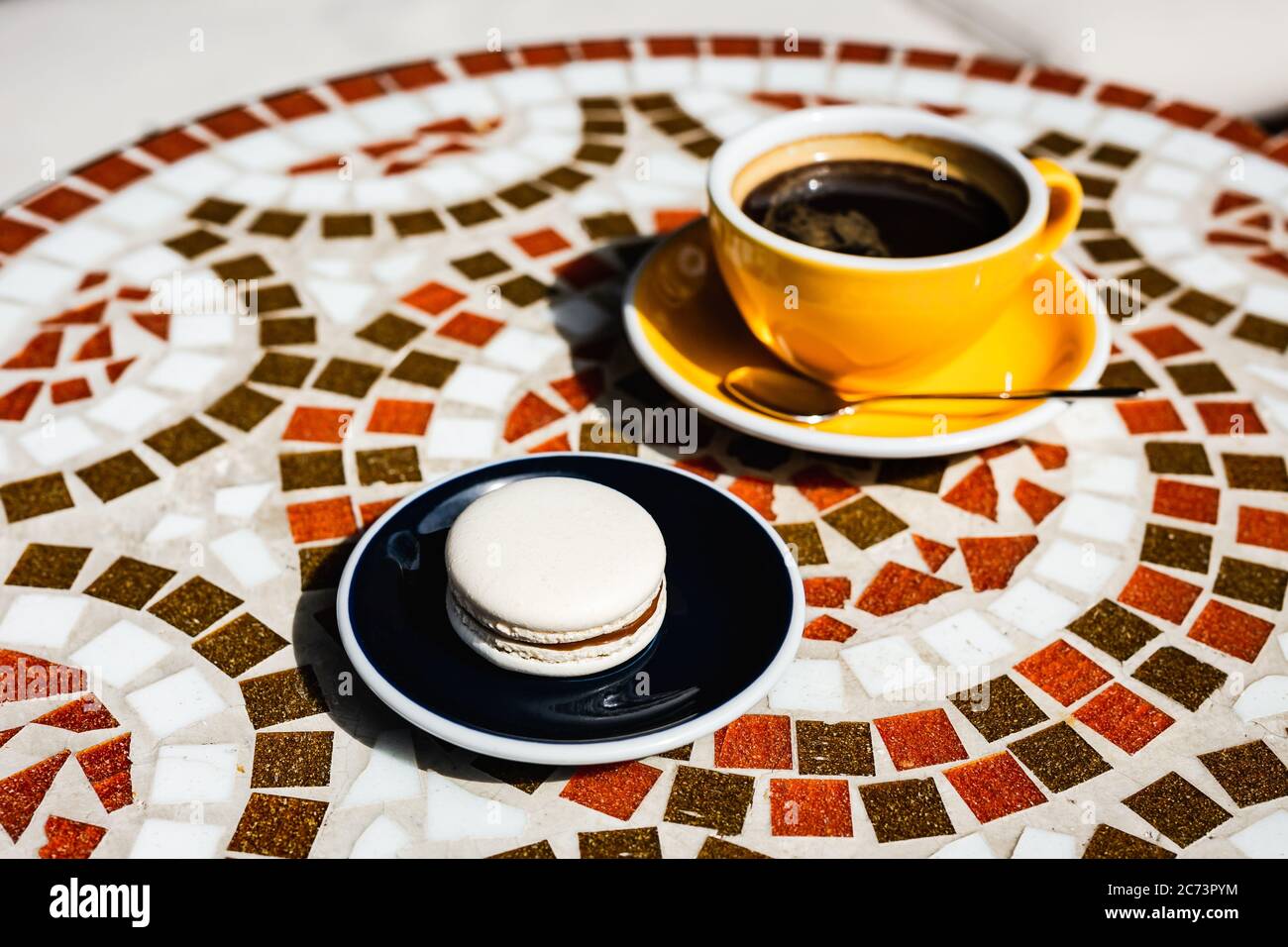 Macaron vanille au chocolat sur une assiette, une tasse jaune de café noir, sur une table en mosaïque de pierre d'un café par une journée ensoleillée Banque D'Images