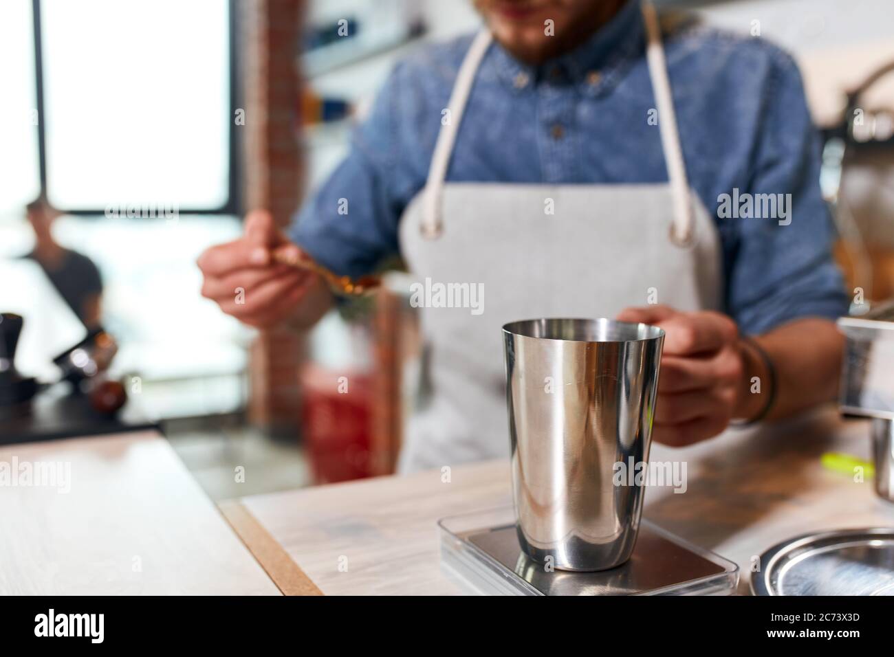 Gros plan d'une cafetière en métal brillant, sur fond flou d'un homme tenant une cuillère argentée avec du café moulu, le barista mesure le café FO Banque D'Images