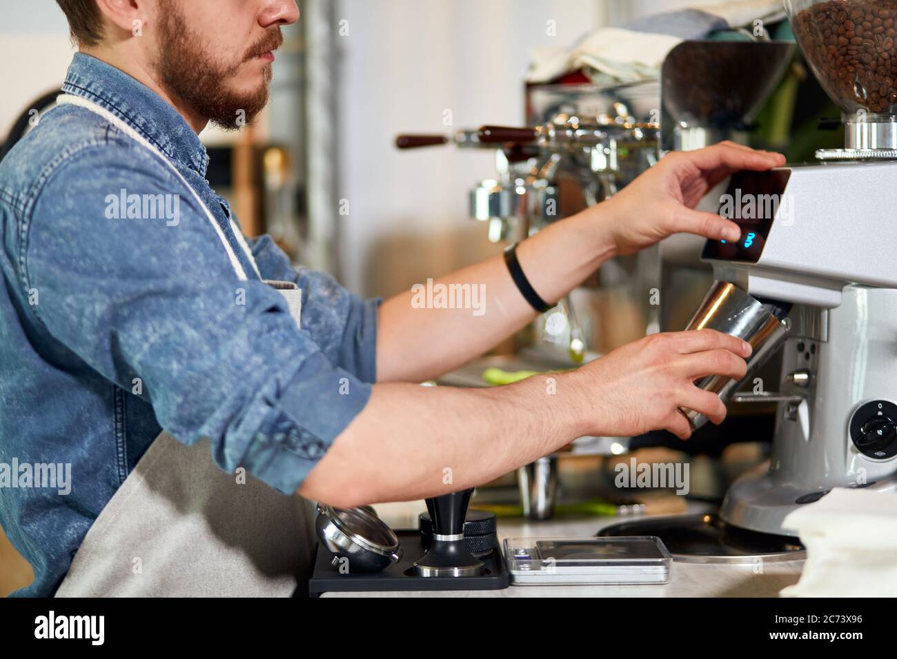 Les mains douces de la barbe le jeune barista tient la cuve en métal, le lait cuit à la vapeur, verse le café moulu, appuie sur le bouton de la machine à café dans le bar, professionnel c Banque D'Images