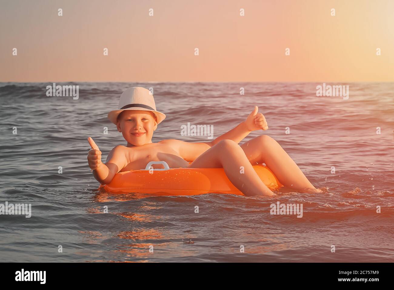 Un garçon caucasien souriant dans un chapeau de paille blanc se détend dans la mer sur un anneau en caoutchouc orange et montre un pouce sur les deux mains dans la soirée au coucher du soleil. Banque D'Images