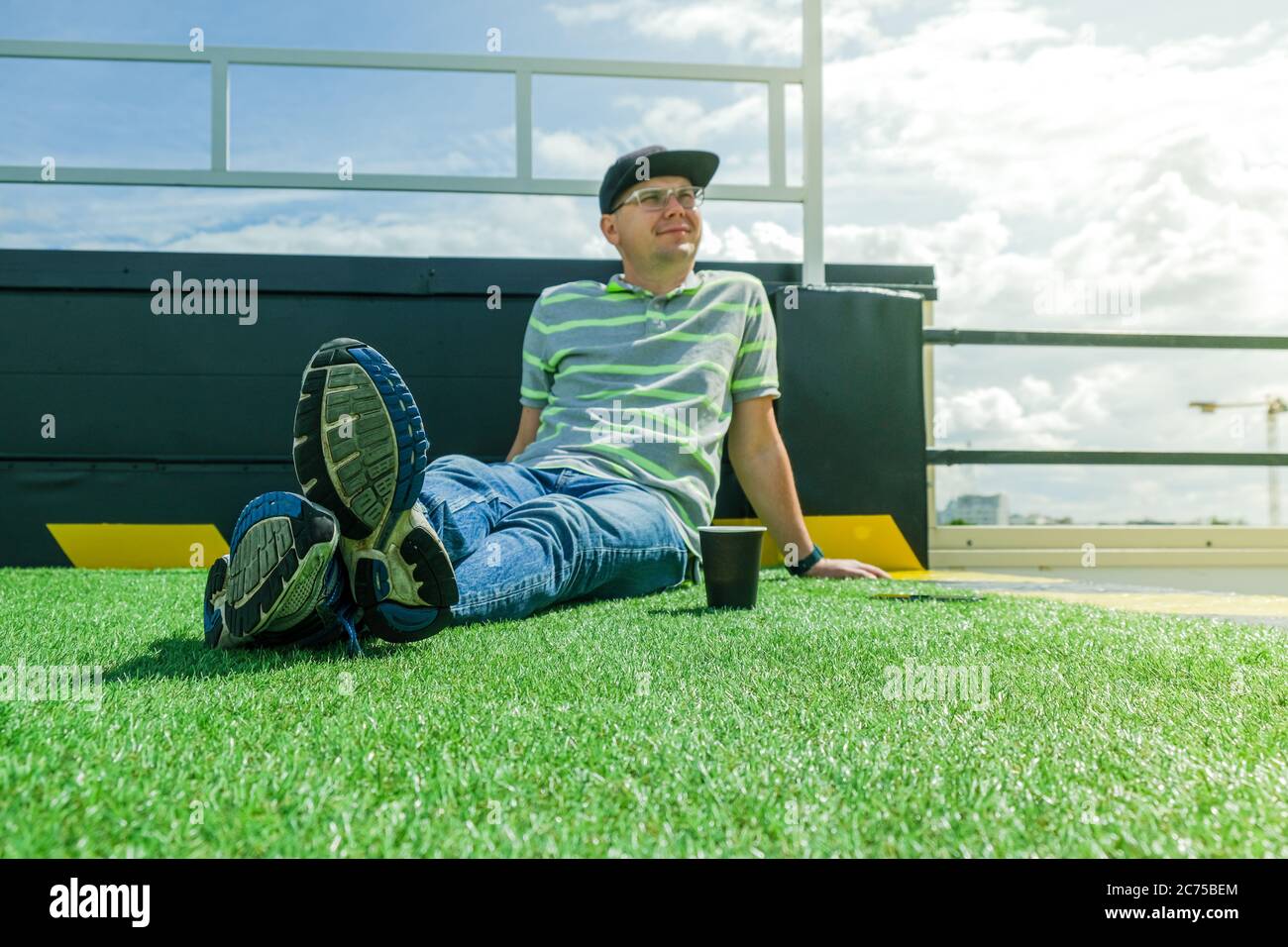 L'homme est assis sur le toit du bâtiment avec une tasse de papier de café et de téléphone cellulaire dans un T-shirt rayé et chapeau noir. Détendez-vous par beau temps. Banque D'Images