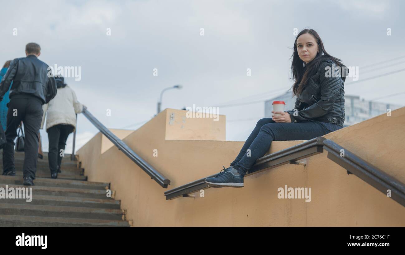 Une jeune femme belle en vêtements décontractés est assise sur une clôture en béton, regarde loin et tient une tasse de café. Banque D'Images
