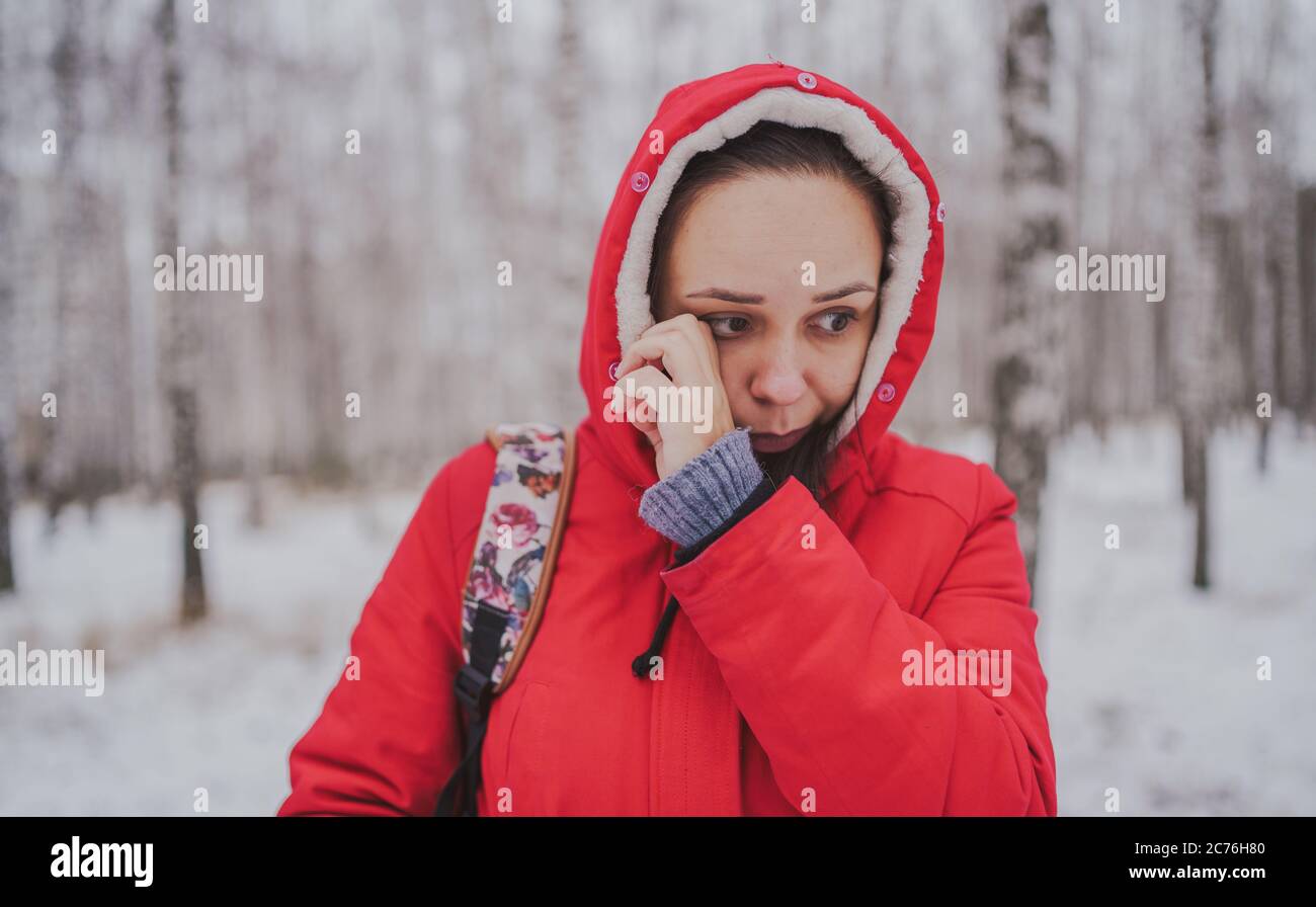 Une jeune femme en blouson rouge et cagoule frotte ses yeux dans la forêt d'hiver. Gros plan. Banque D'Images